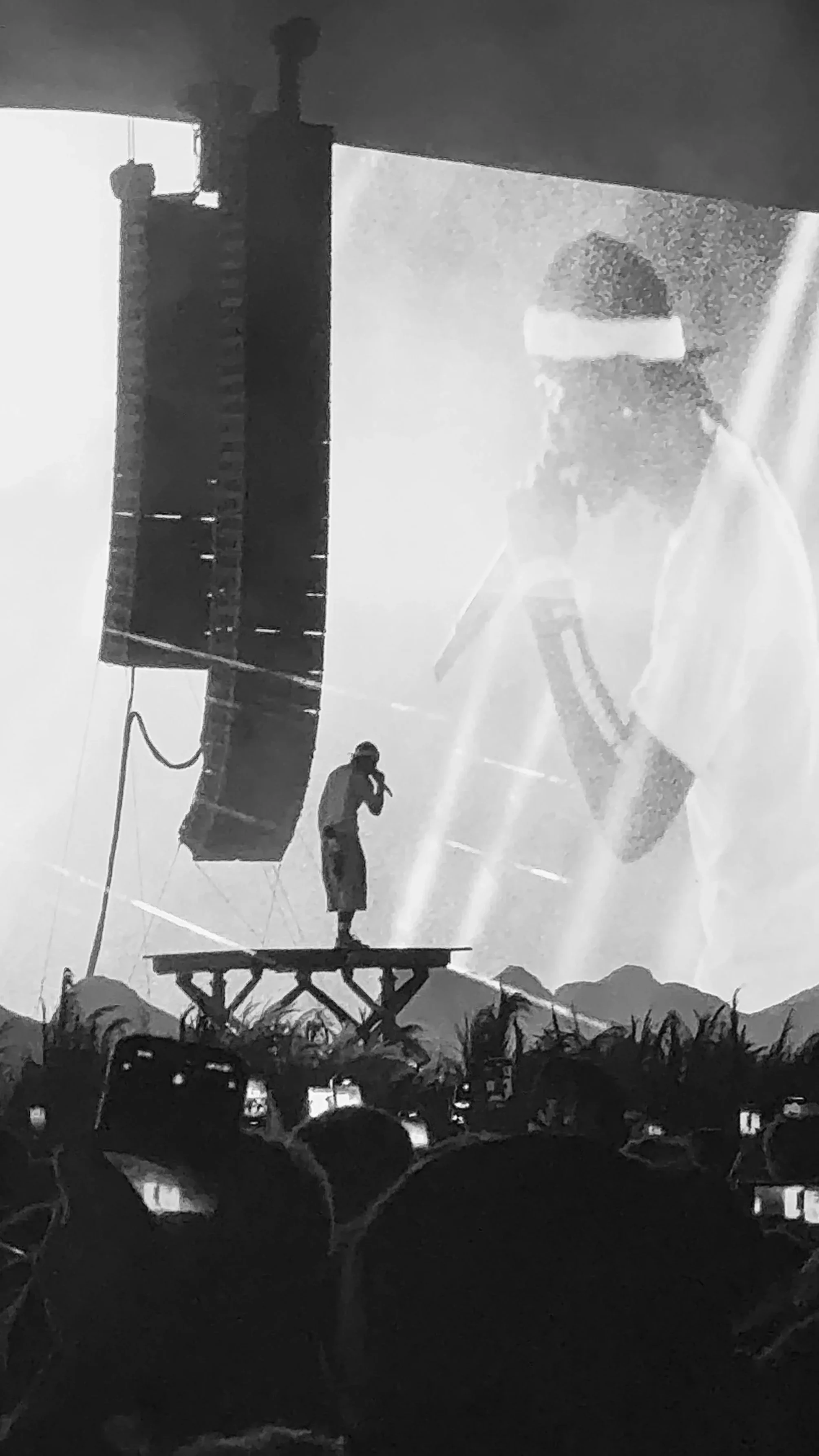 A singer stands on a small platform holding a microphone on stage, with a large screen behind them displaying their live image, and crowd members capturing the moment with their phones. Travis Scott Leeds Festival