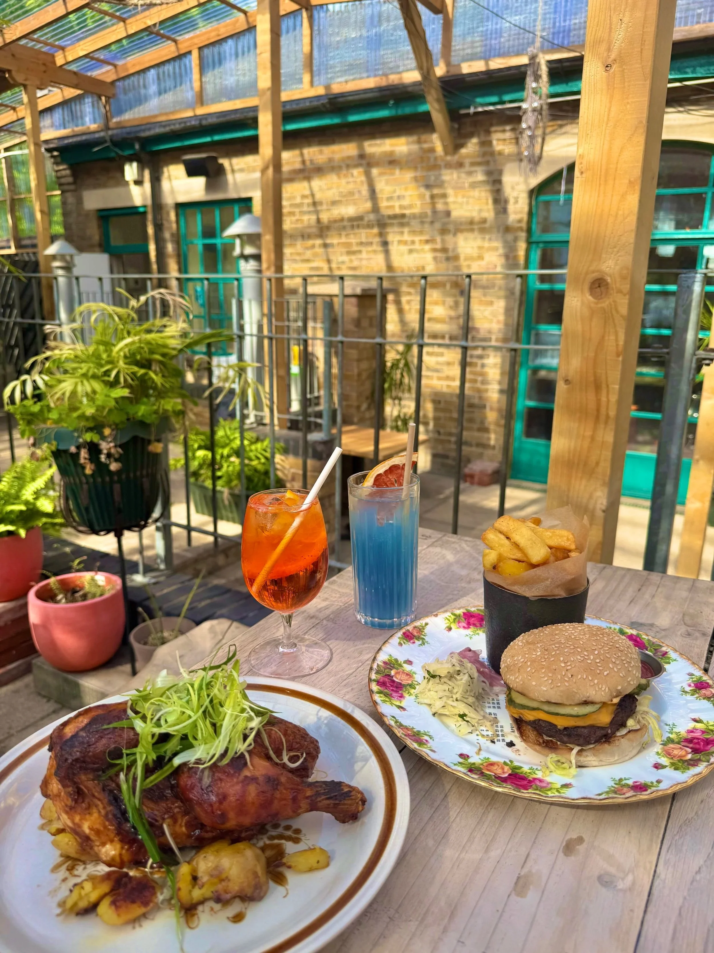 Outdoor dining table with a roasted chicken garnished with greens, a plate with a cheeseburger with fries, two colorful cocktails, and potted plants in the background.