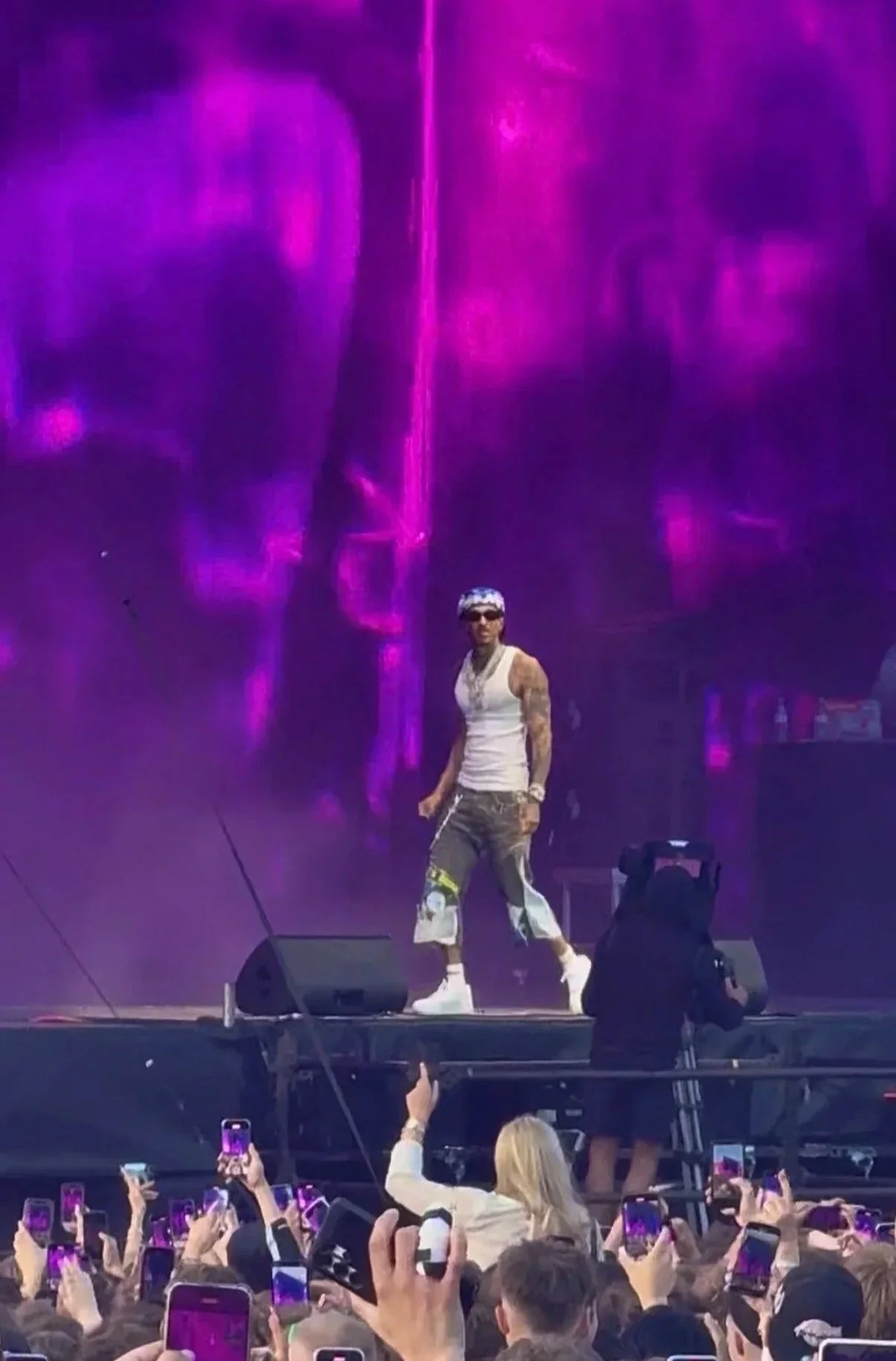 A male performer on stage with a purple and pink light display behind him, wearing sunglasses, a sleeveless white top, colorful pants, and white shoes, standing in front of a crowd taking photos.