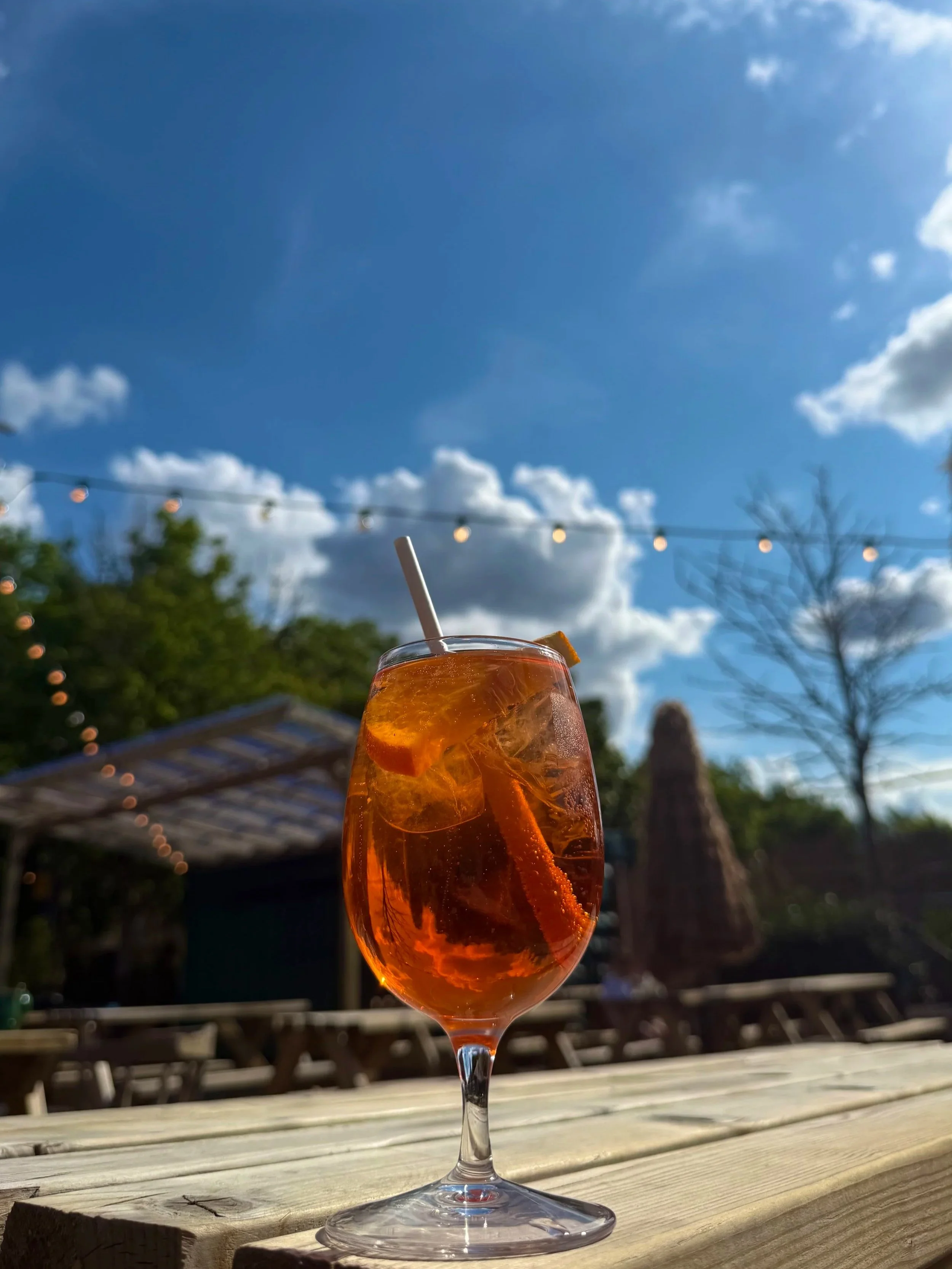 A glass of iced orange-colored drink with a straw on a wooden table outdoors, with a blue sky and clouds in the background.