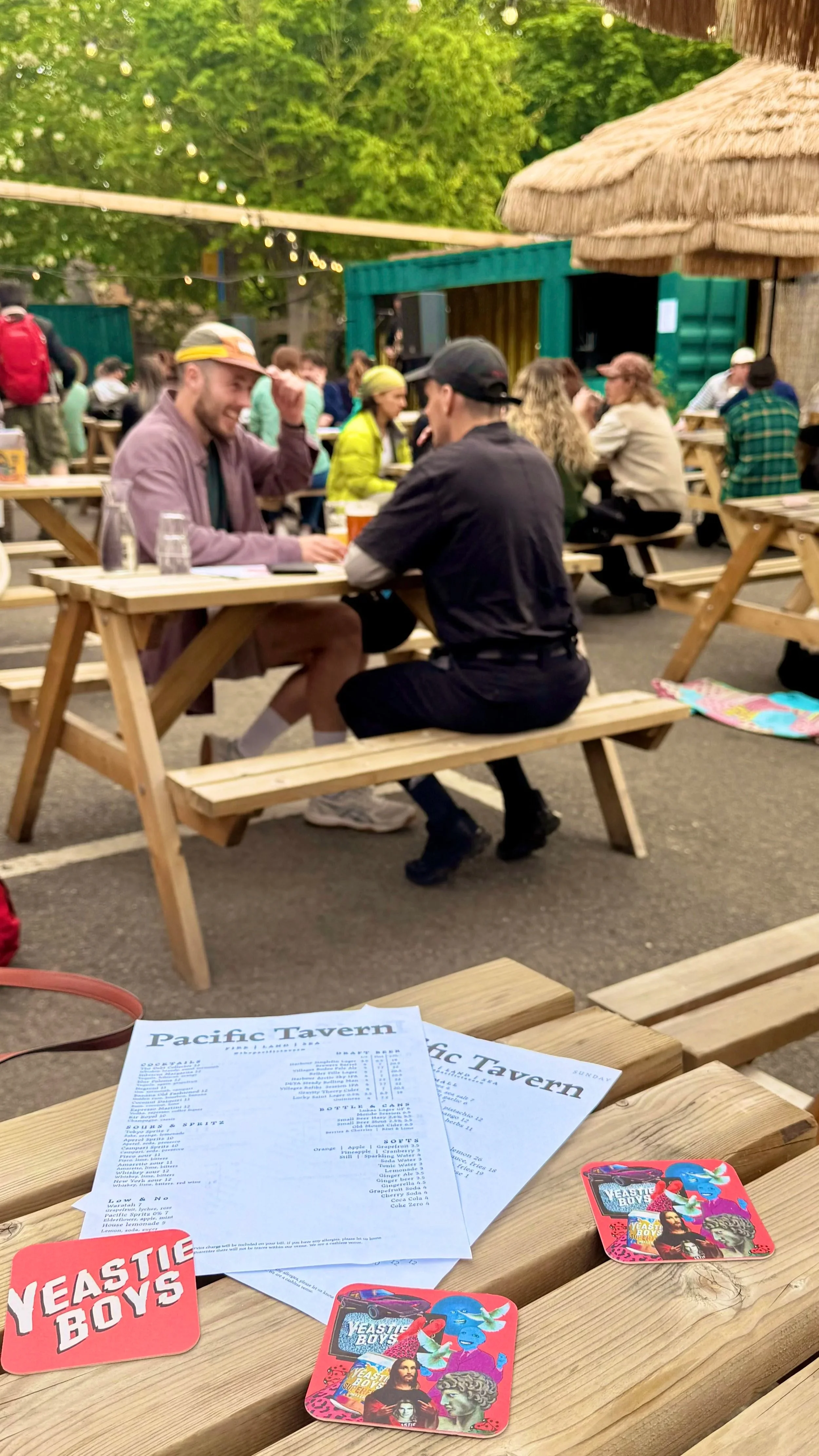People enjoying drinks at outdoor picnic tables at a lively bar or brewery with string lights and green trees in the background. Men and women are socializing, and there are menus and coasters on the wooden table in the foreground.
