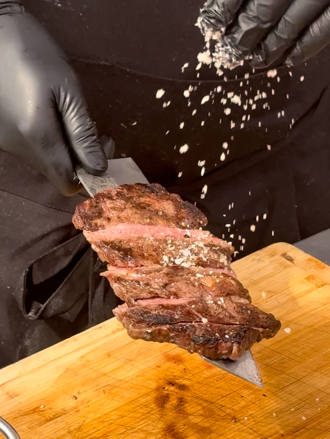 A person wearing black gloves slices a cooked steak on a wooden cutting board, seasoning falling from their hand.