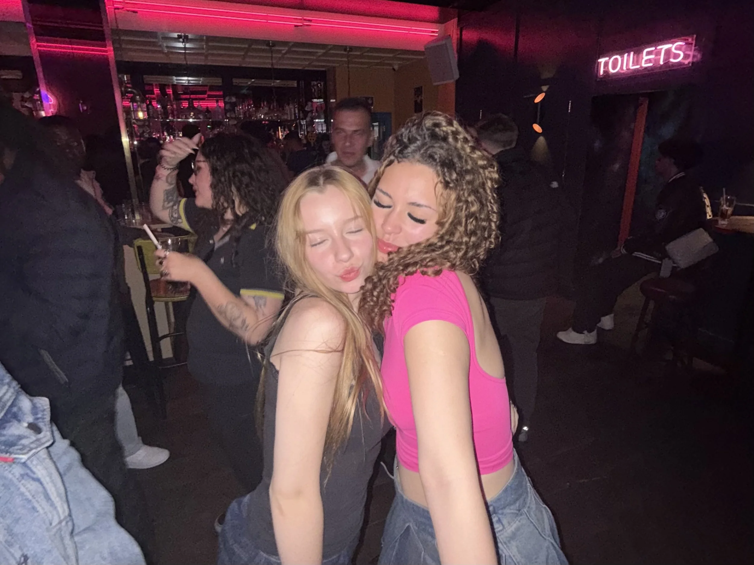 Two young women, one with blonde hair and the other with curly brown hair, are close together, cheeks touching, with their eyes closed and lips puckered, in a nightclub with a pink neon sign that says 'TOILETS' in the background. Simmons