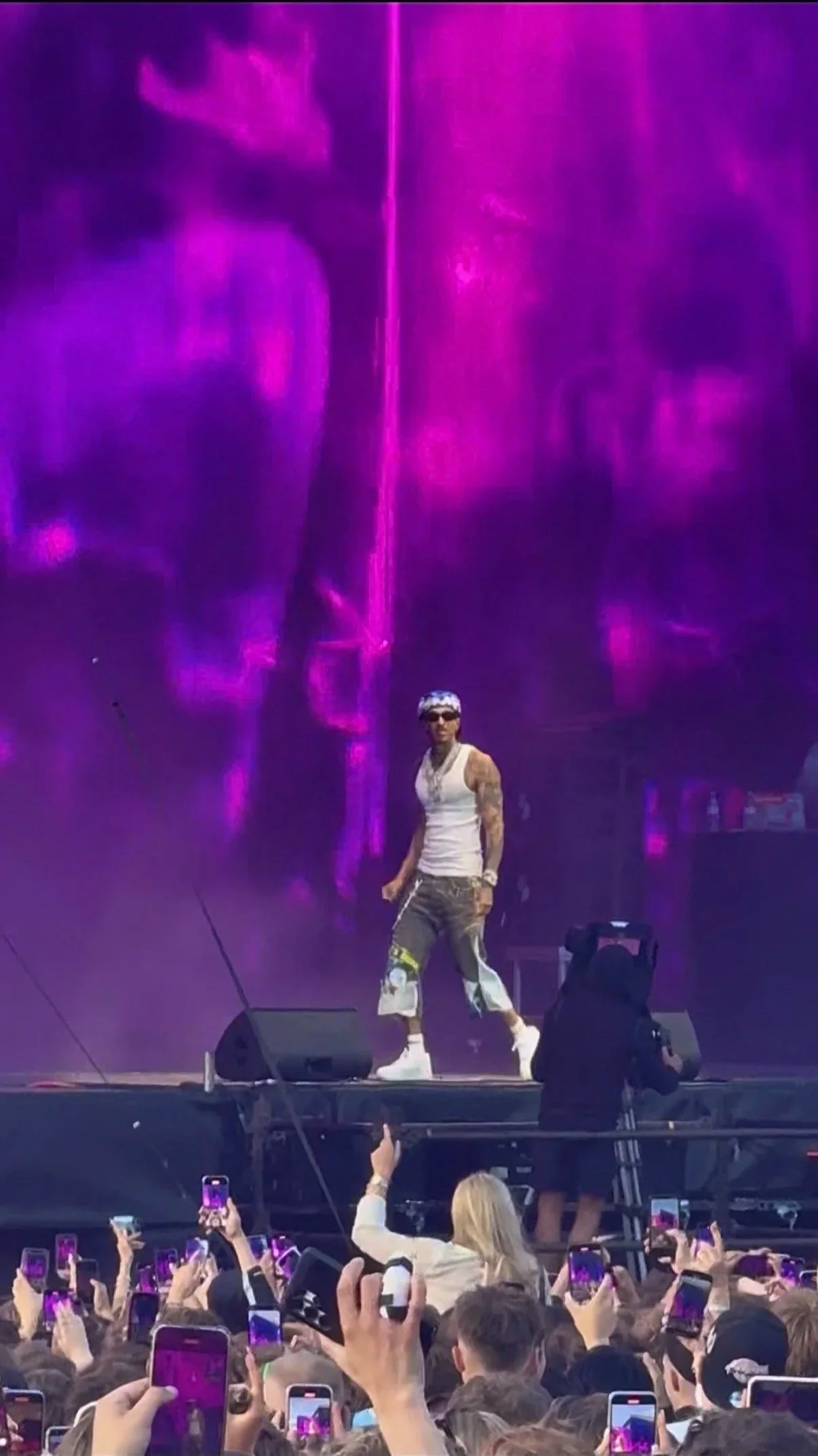 A performer on stage with purple lighting and fog, wearing a white tank top, baggy pants with patches, sunglasses, and a bandana, in front of a crowd taking photos. D-Block Europe Young Adz Leeds Festival