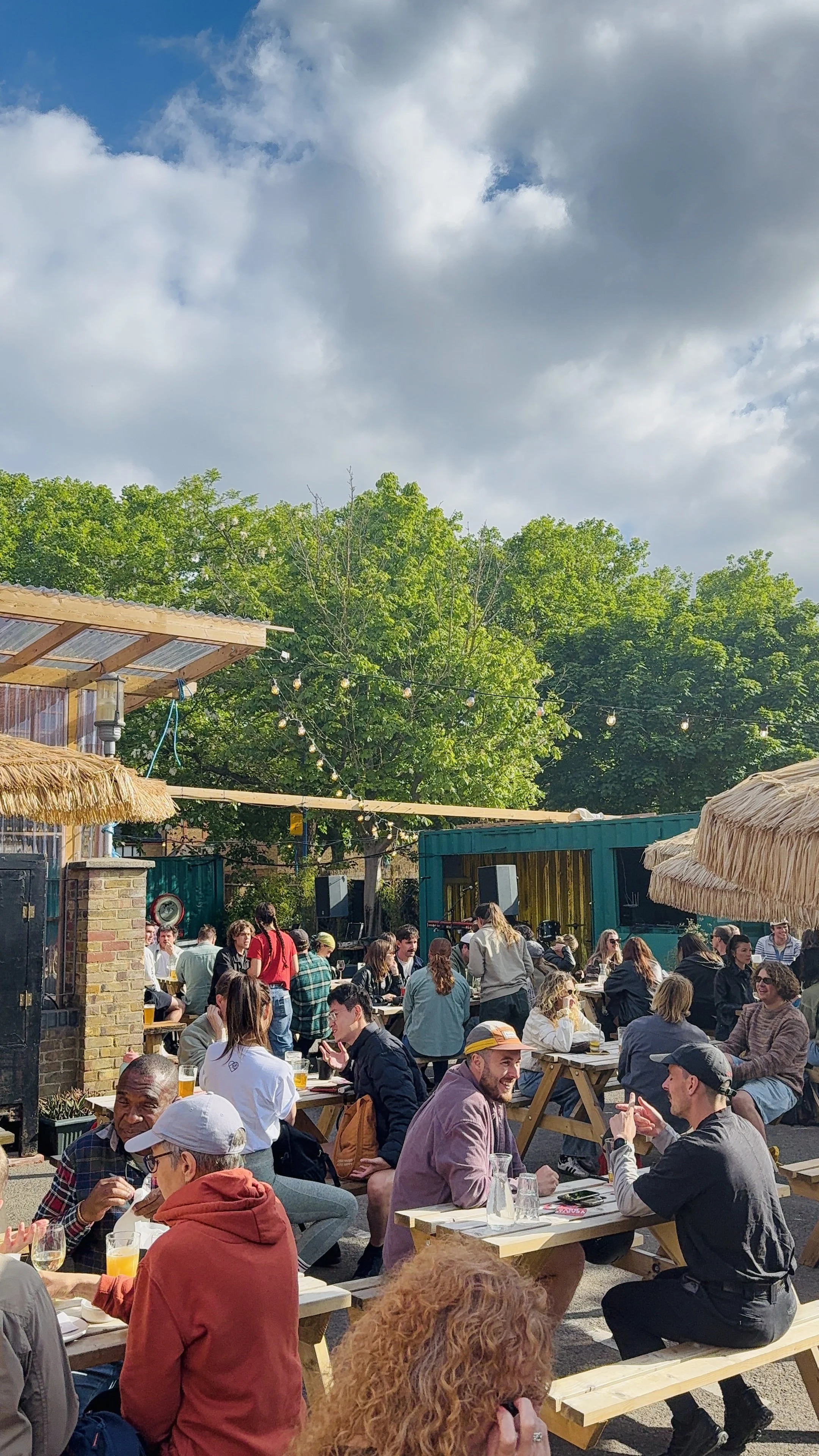 People dining outdoors at a social gathering or event under string lights, with trees in the background and a partly cloudy sky.
