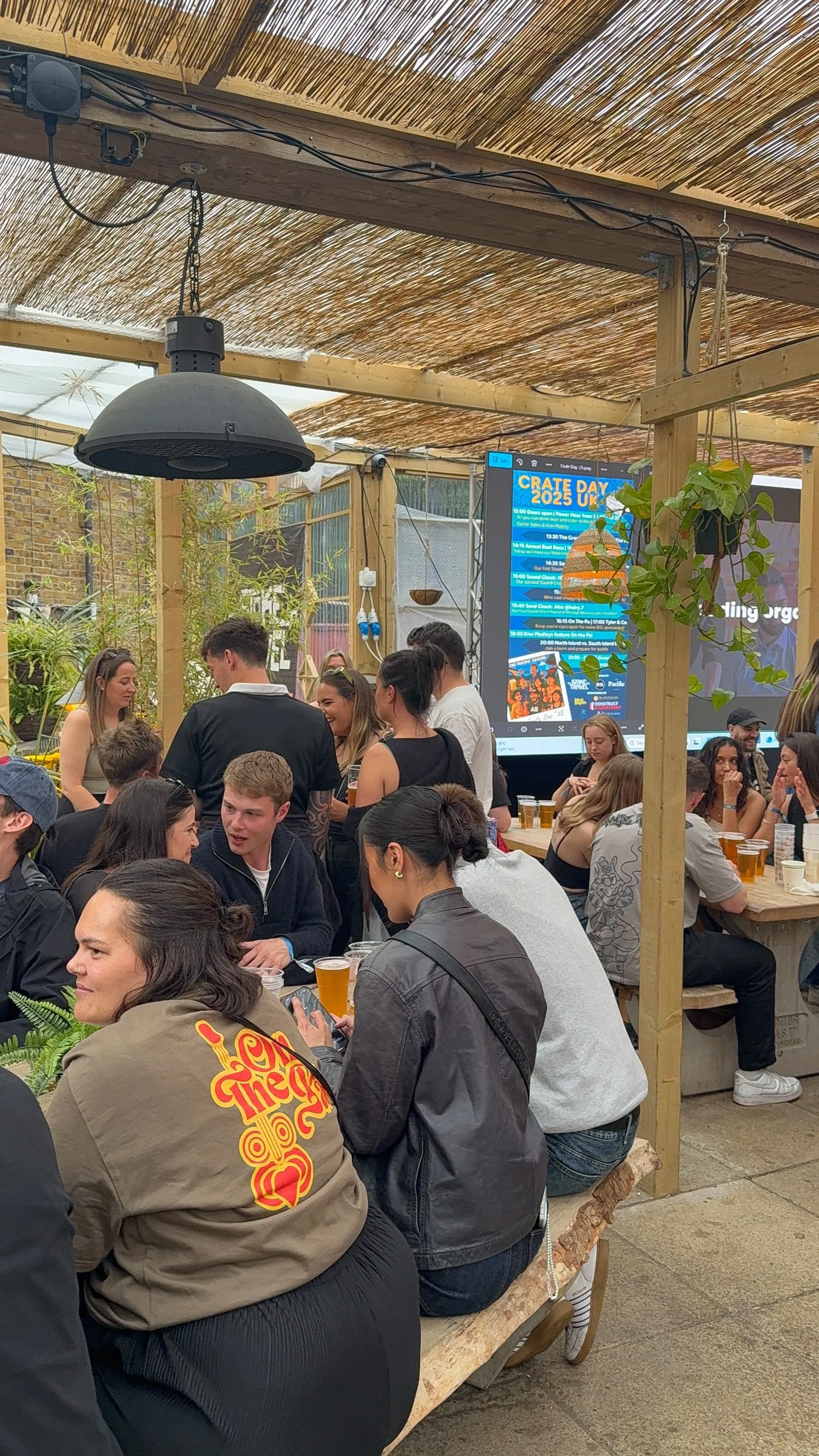 People gathered in a covered outdoor space with wooden beams and a thatched roof, socializing and drinking beer, with a large digital screen displaying 'Crate Day 2025 UK' event schedule.