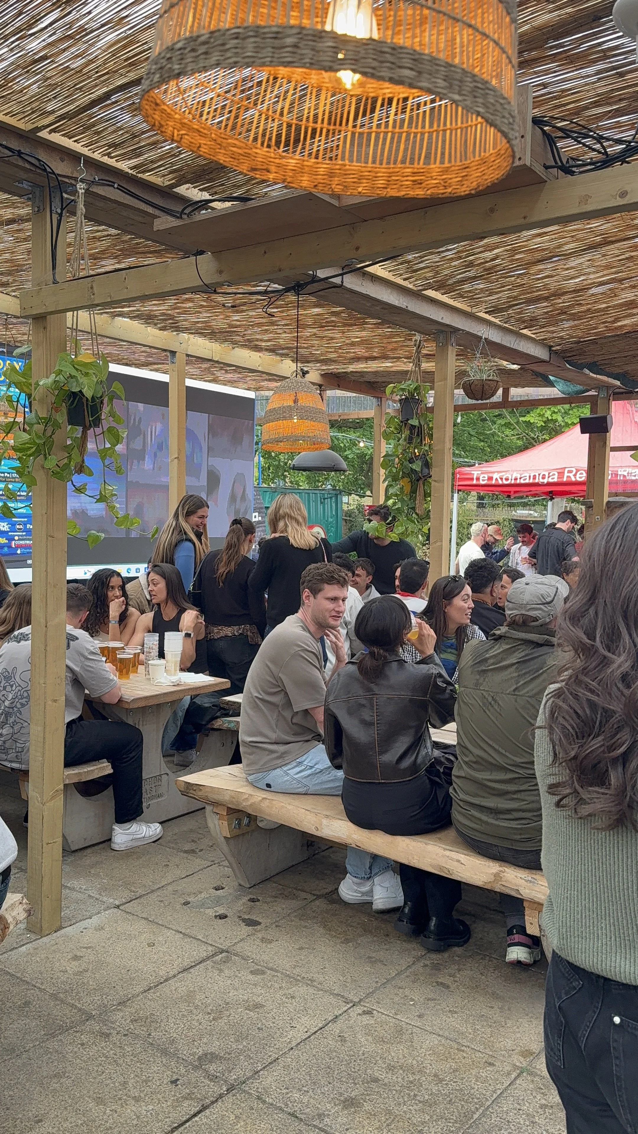 People sitting and socializing at outdoor wooden picnic tables under a thatched roof at a social gathering or event.