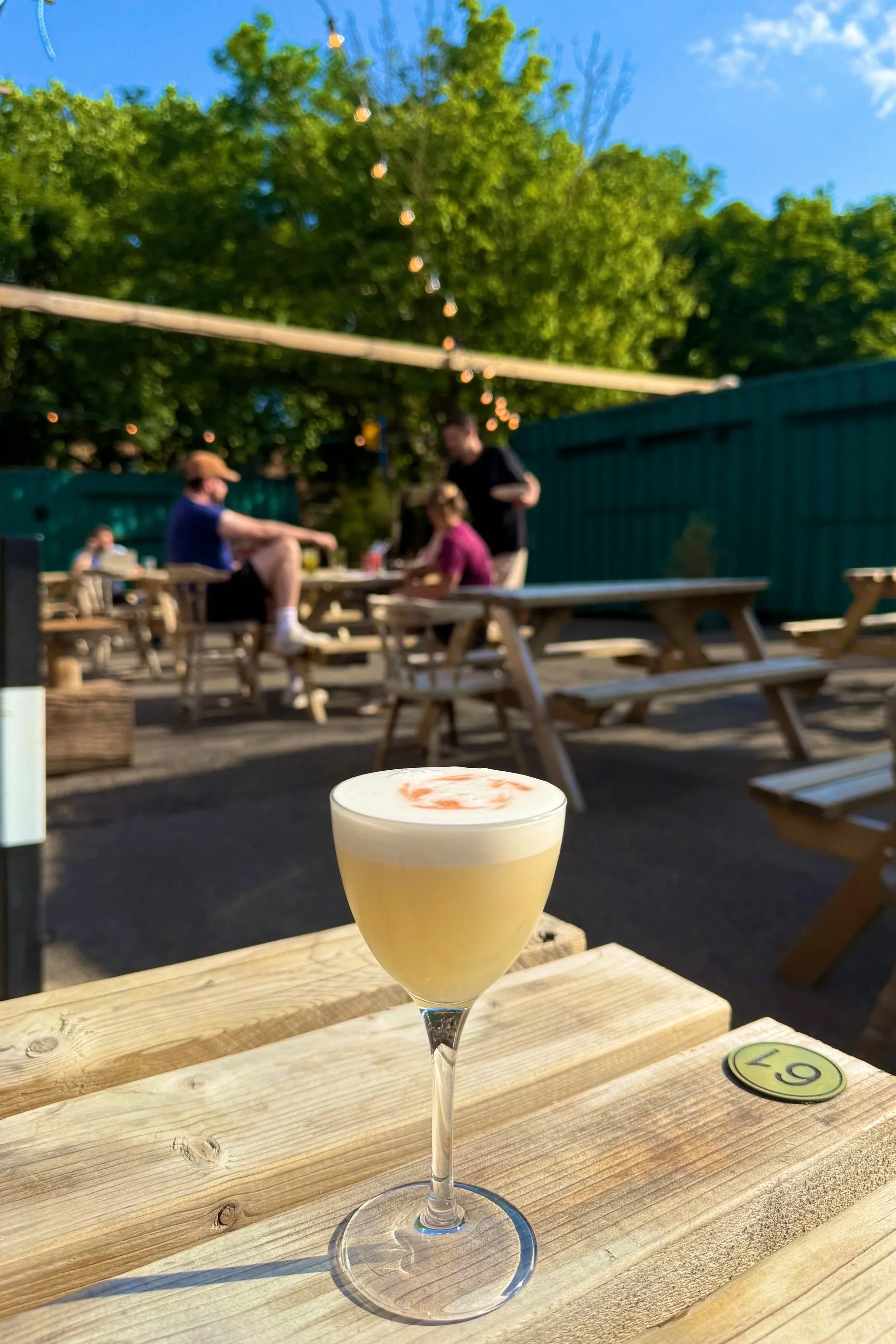 A cocktail with a frothy top and orange swirl, placed on a wooden table in an outdoor seating area of a restaurant or bar, with picnic tables, string lights, and trees in the background.
