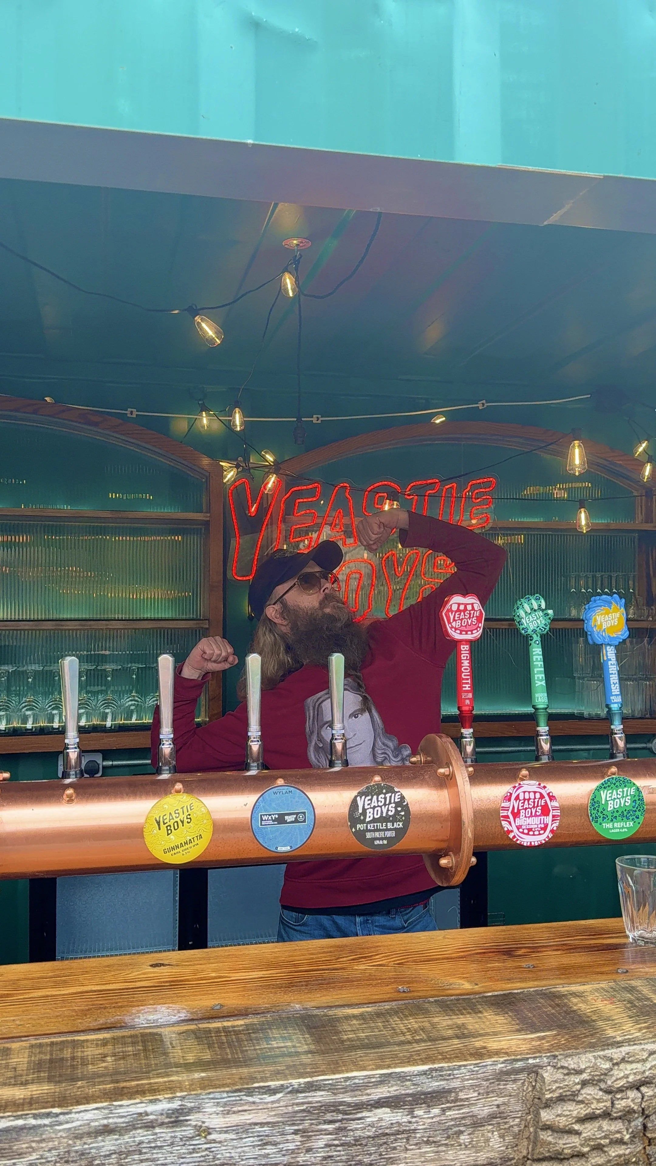A man with long hair, a beard, sunglasses, and a cap flexing his arm muscles behind a bar with a copper pipe beer dispenser at Yeaste Boys brewery, with colorful taps and a neon sign in the background.