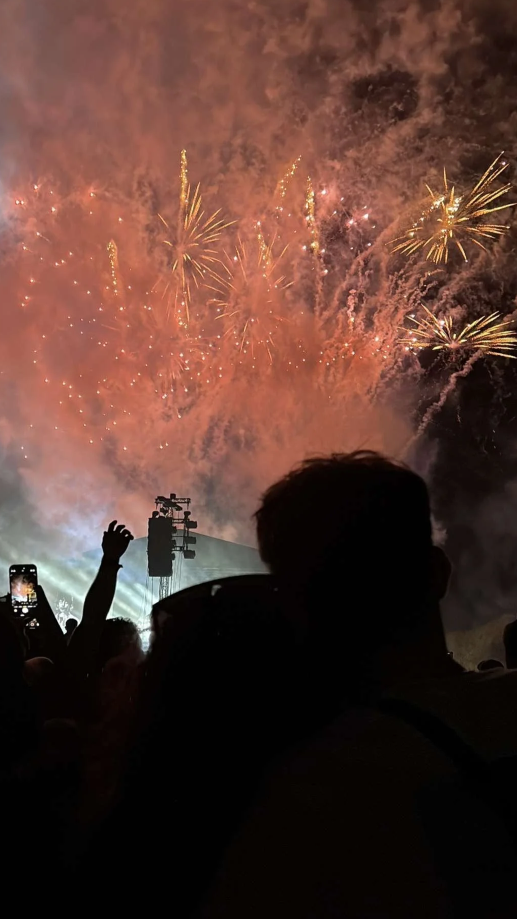 Fireworks display illuminating the night sky with silhouettes of people watching and capturing the event on their phones.