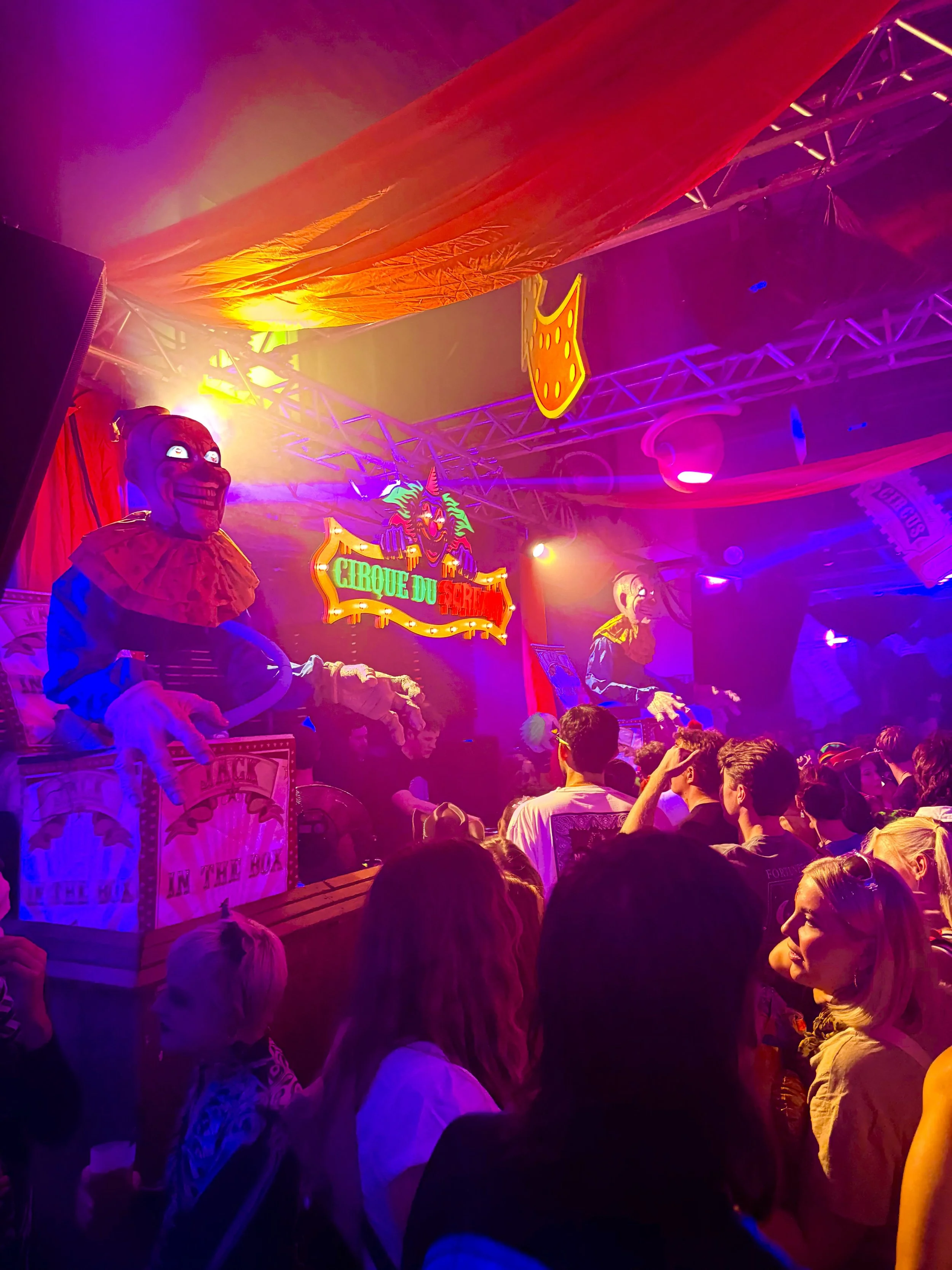 A crowded circus tent with colorful lighting and circus props. Clowns with masks and costumes are visible, along with a sign that reads 'Cirque du' and a large clown face decoration.