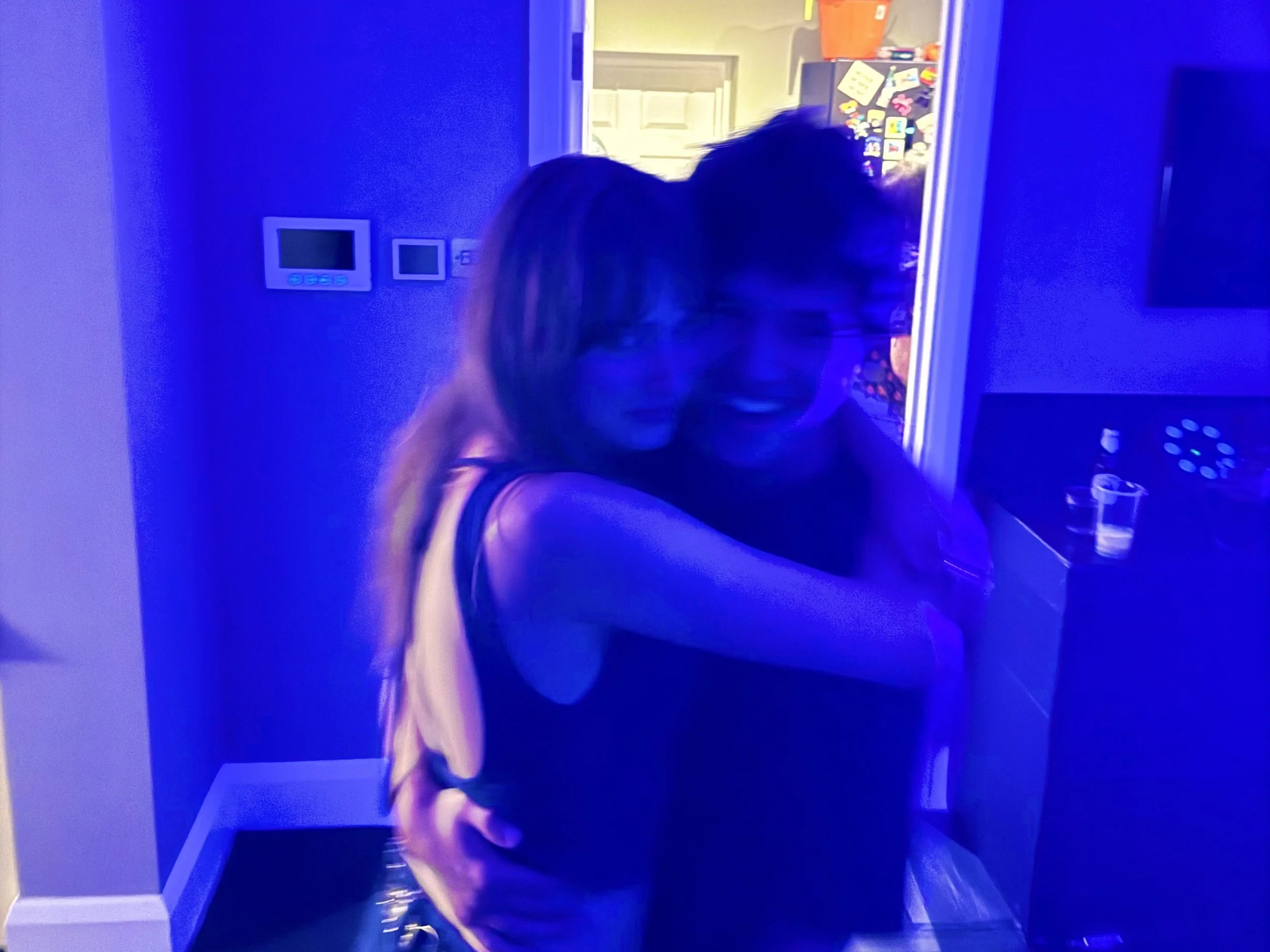 A woman with long hair, wearing a pink top, is taking a selfie in a room illuminated by blue light. The room has a door, wall-mounted control panels, a counter with various items, and a glasses of water visible nearby.