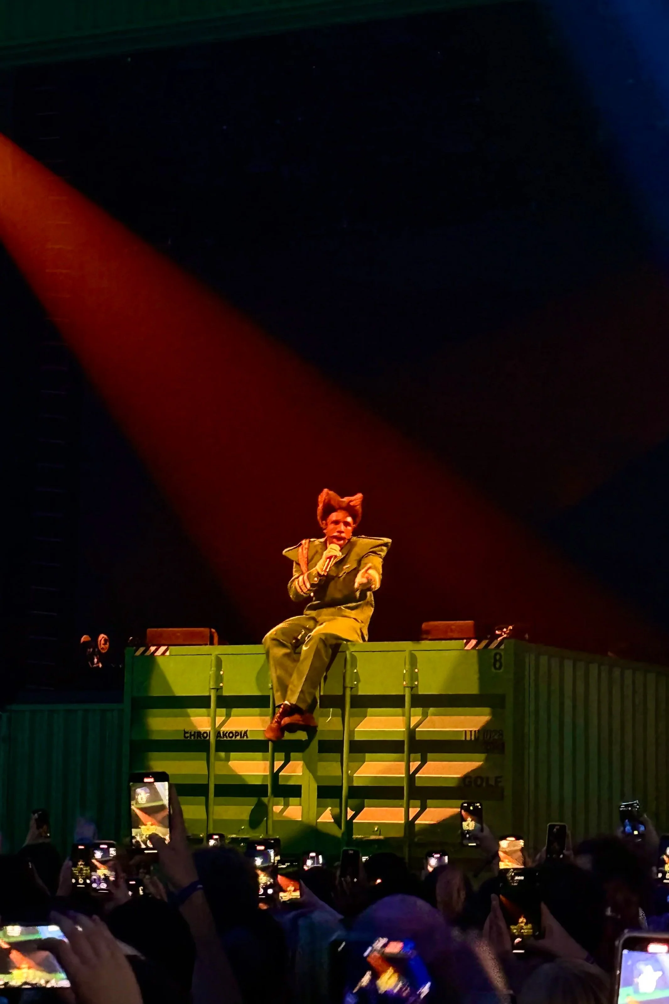 Performer dressed in green military-like costume sitting on top of a green shipping container on stage, holding a microphone and speaking, with an audience capturing photo. Tyler, the Creator O2 Arena London