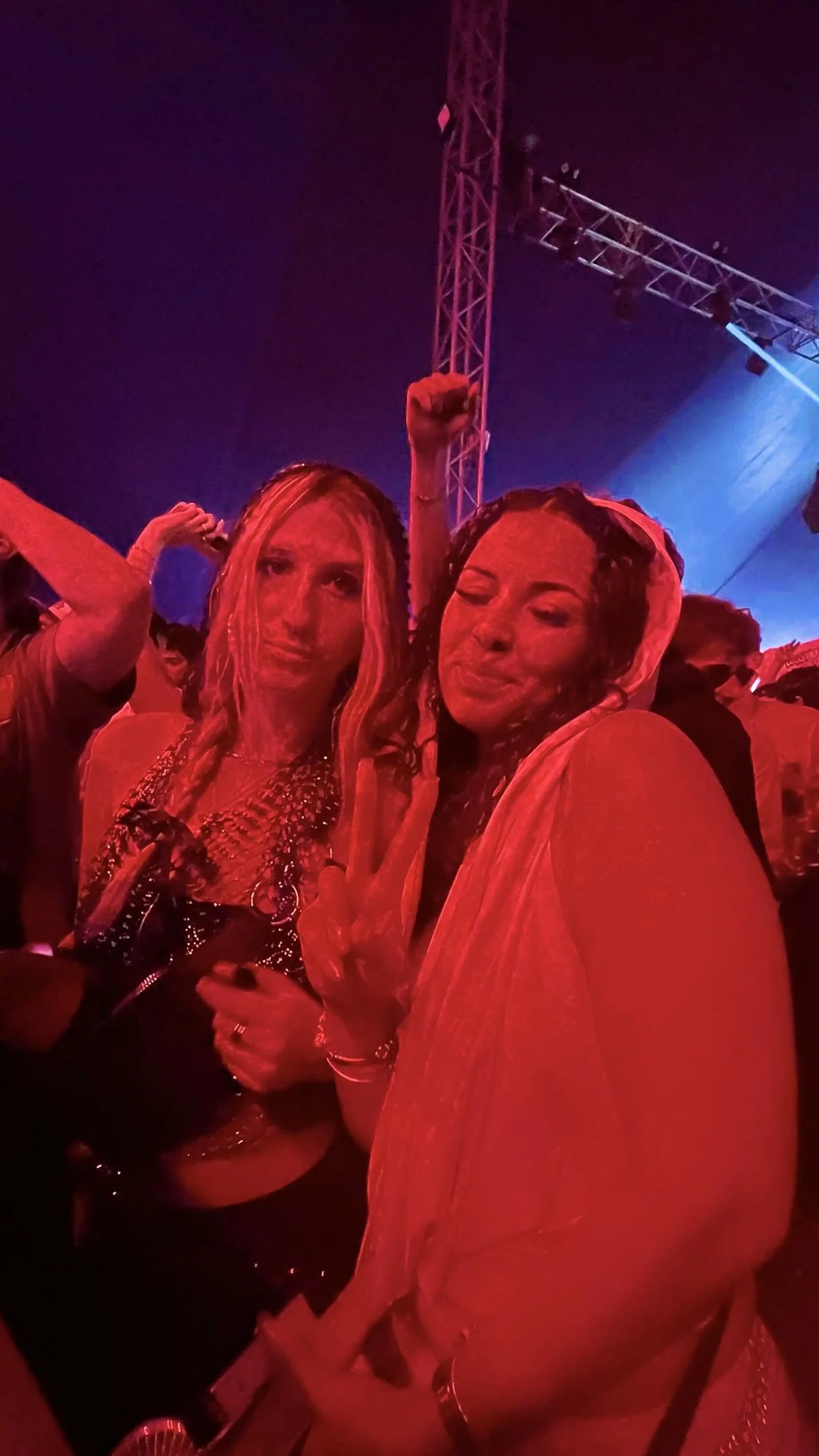 Two women at a concert or nightclub, one making a peace sign and the other raising her fist, under colorful stage lighting.