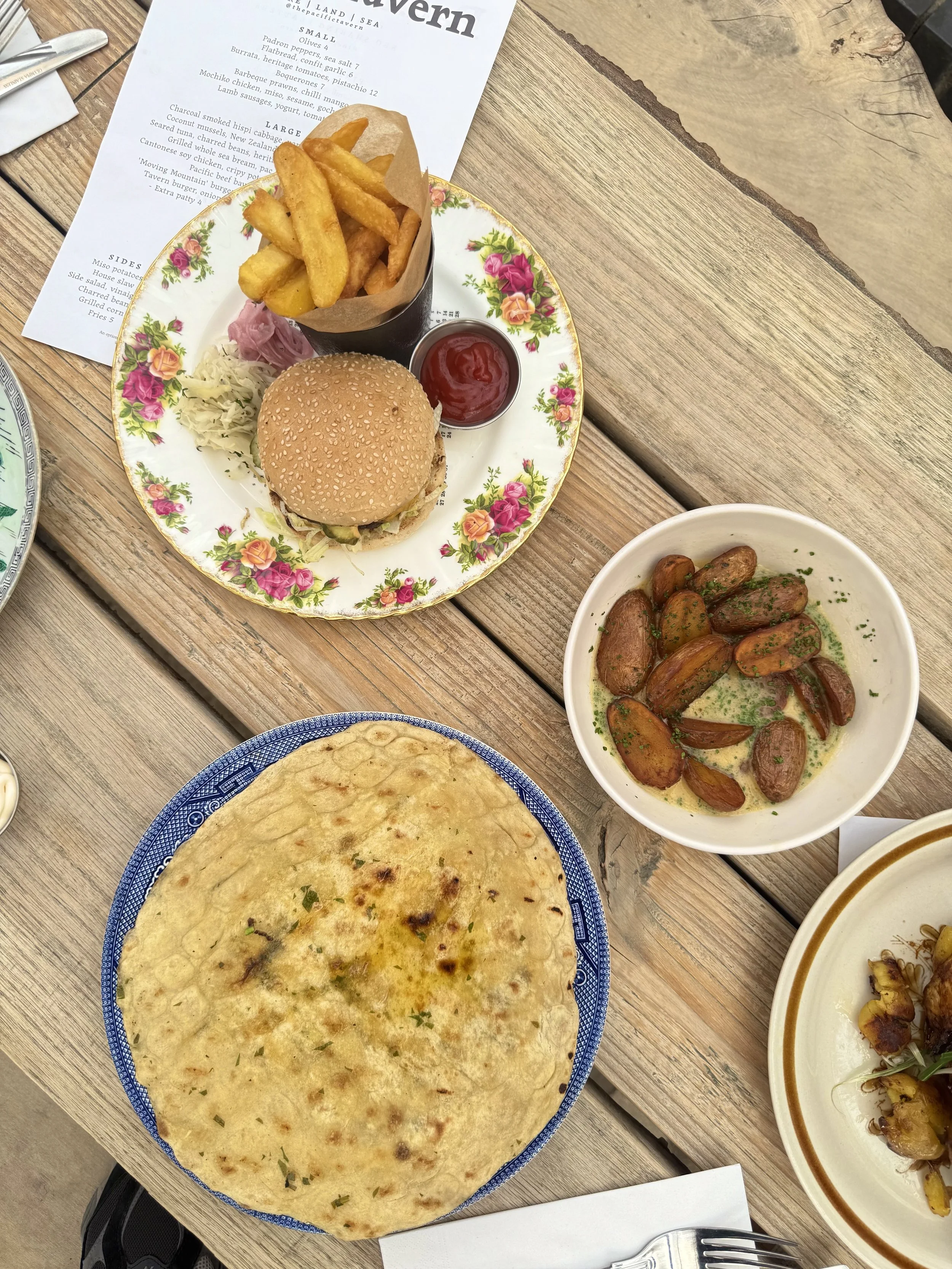 A meal on a wooden table includes a plate with flatbread, a burger with fries and ketchup, a bowl of roasted garlic potatoes, and a plate with grilled skewers.