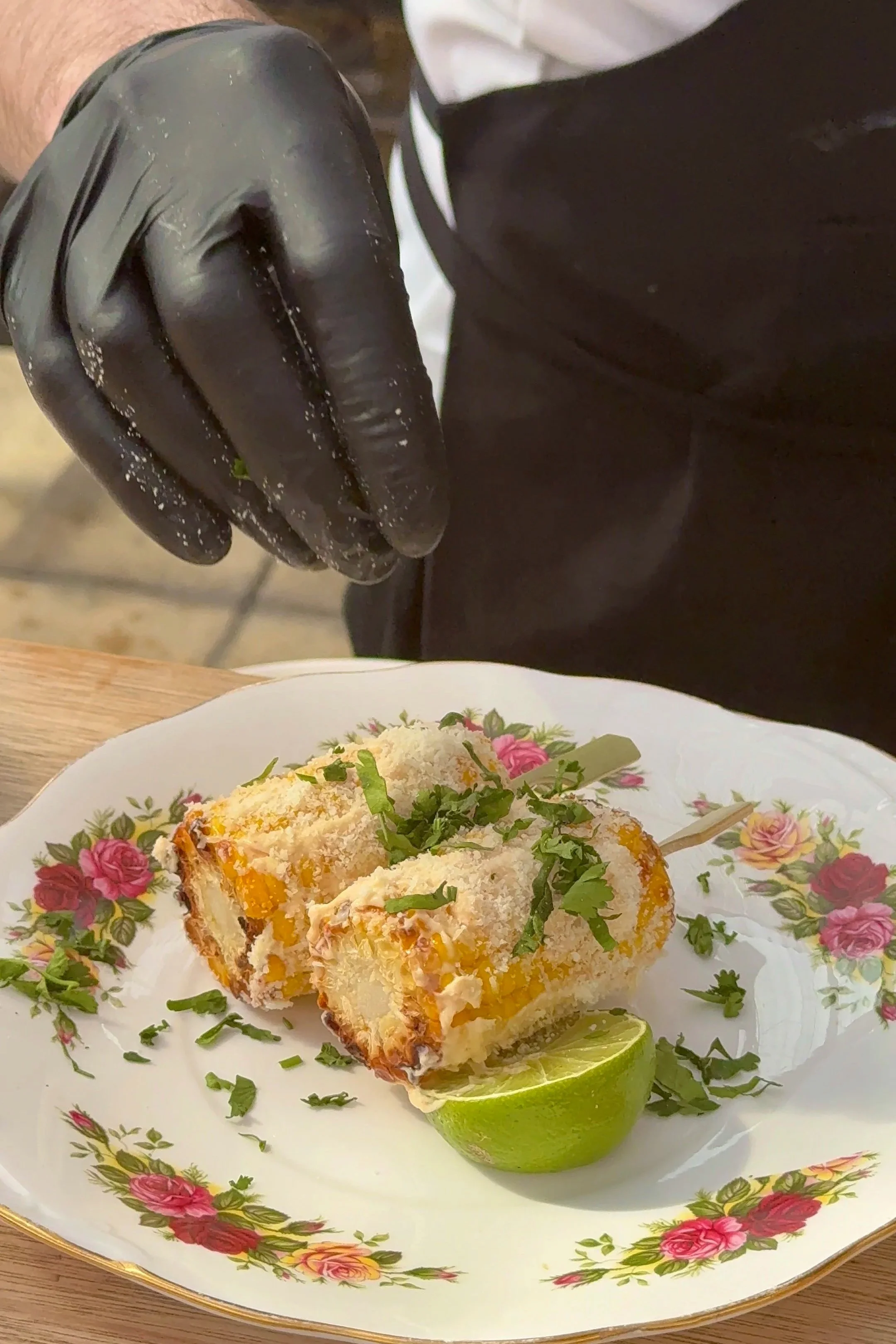 Chef garnishing breaded stuffed corn on the cob with cilantro, served on a floral china plate with a lime wedge.