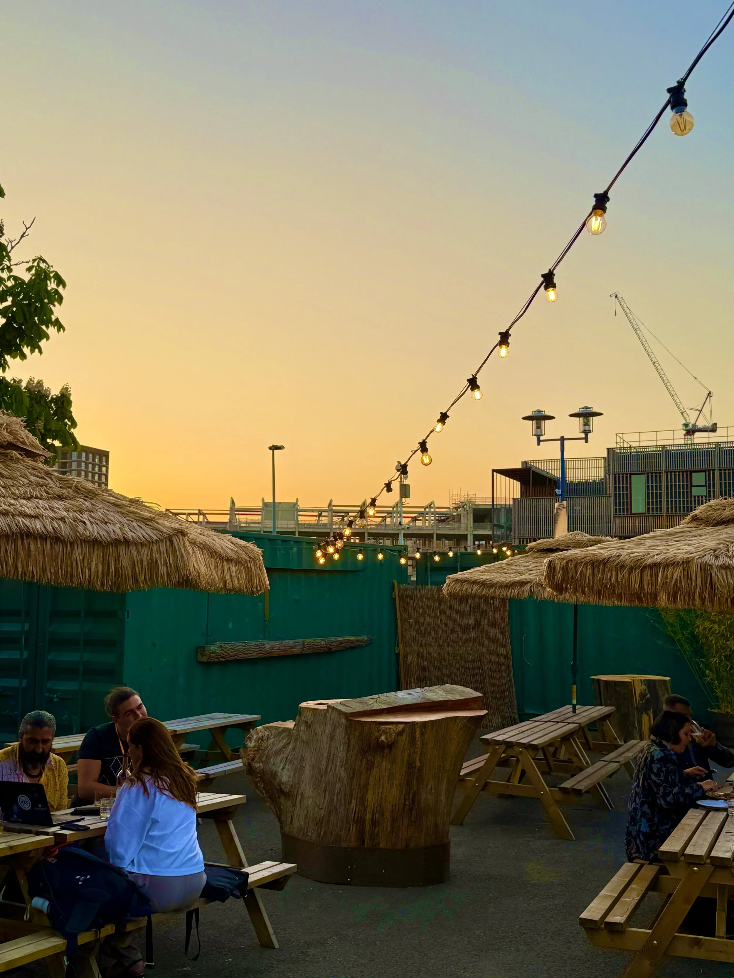 Outdoor patio with wooden tables and benches, some people sitting and talking, string lights hanging above, thatched roof awnings, green fence, sky with sunset colors, construction cranes in the background.