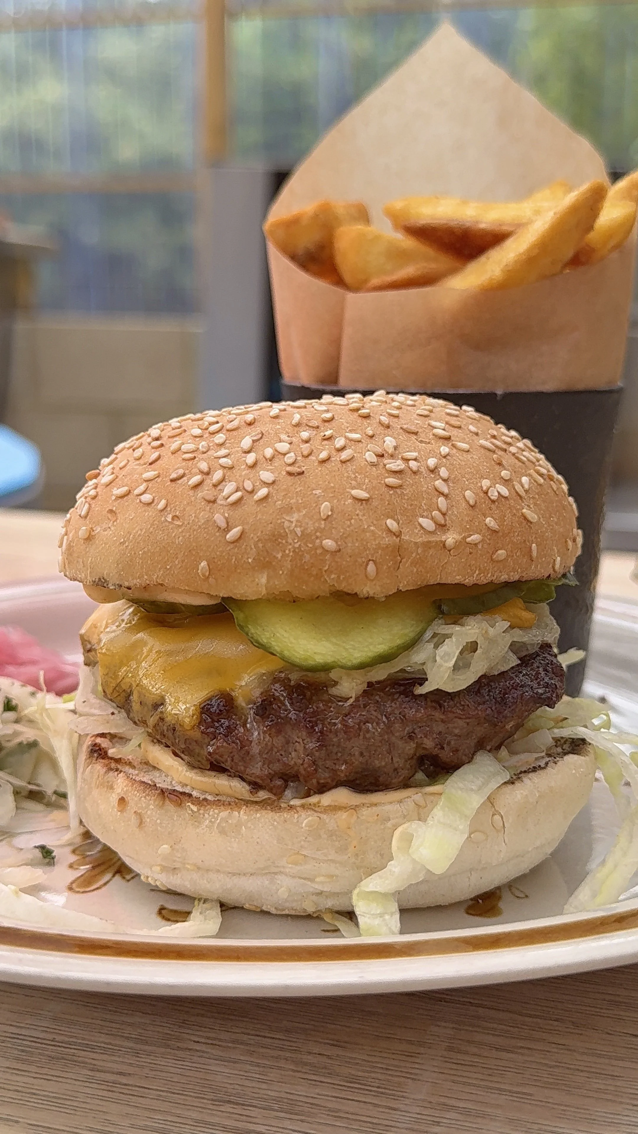 Burger with lettuce, pickles, cheese, and a sesame seed bun, served with a side of French fries in a paper cone.