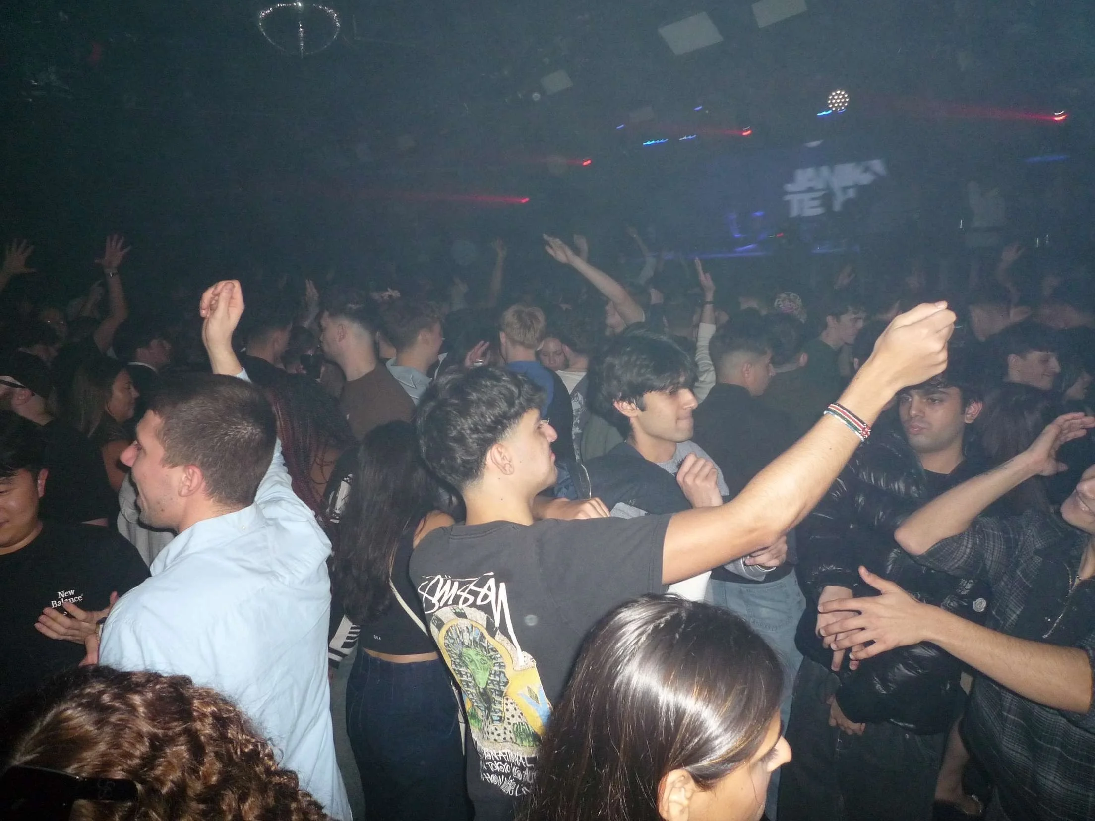 A crowded dance floor at a nightclub with many young people dancing and enjoying themselves. People are raising their hands, talking, and having fun amidst colorful lights and a foggy atmosphere. Ministry Of Sound