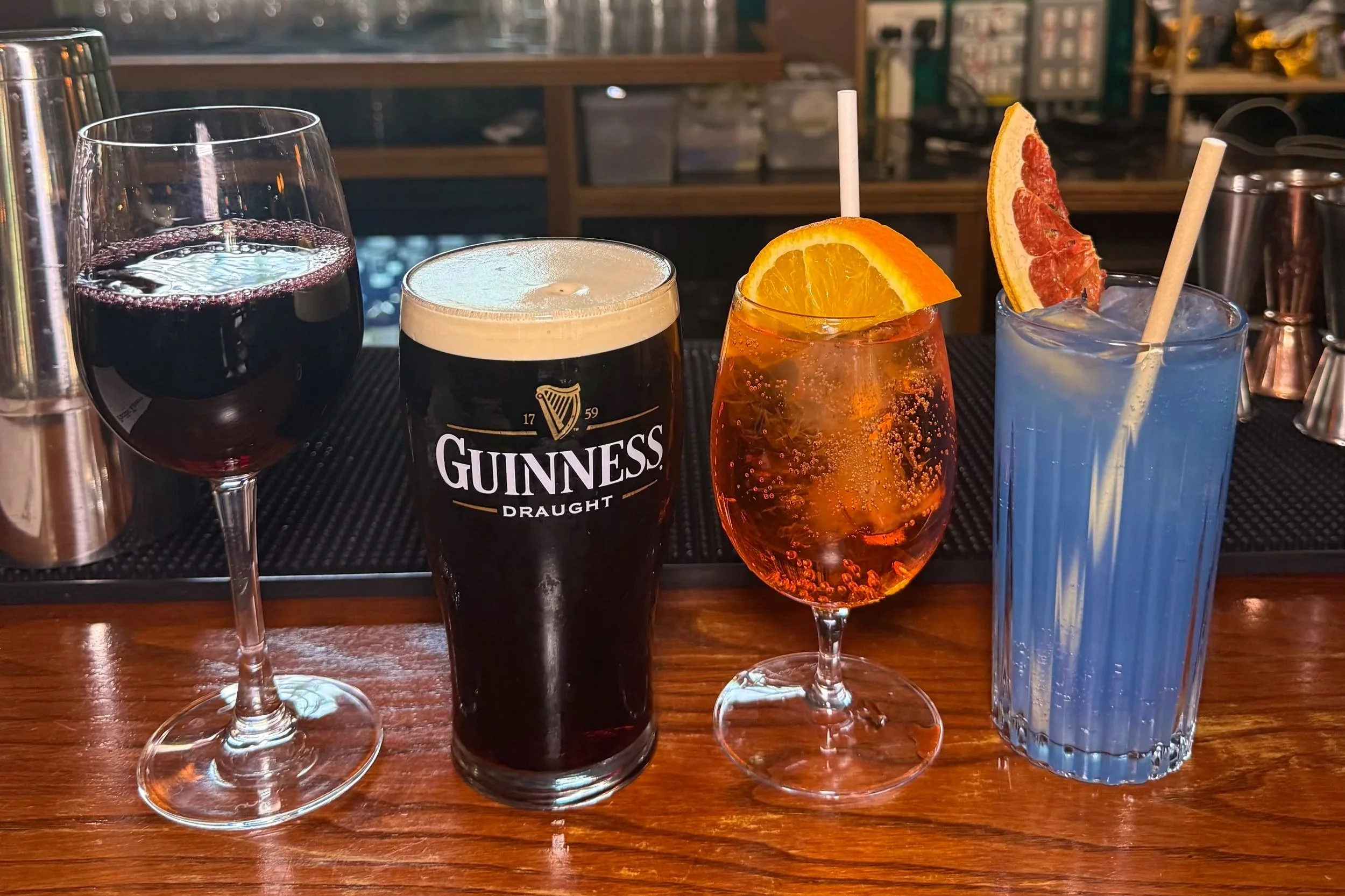 A row of four different drinks on a bar counter. From left to right: a glass of red wine, a Guinness beer, a cocktail with orange and grapefruit slices, and a blue cocktail with a lemon straw.