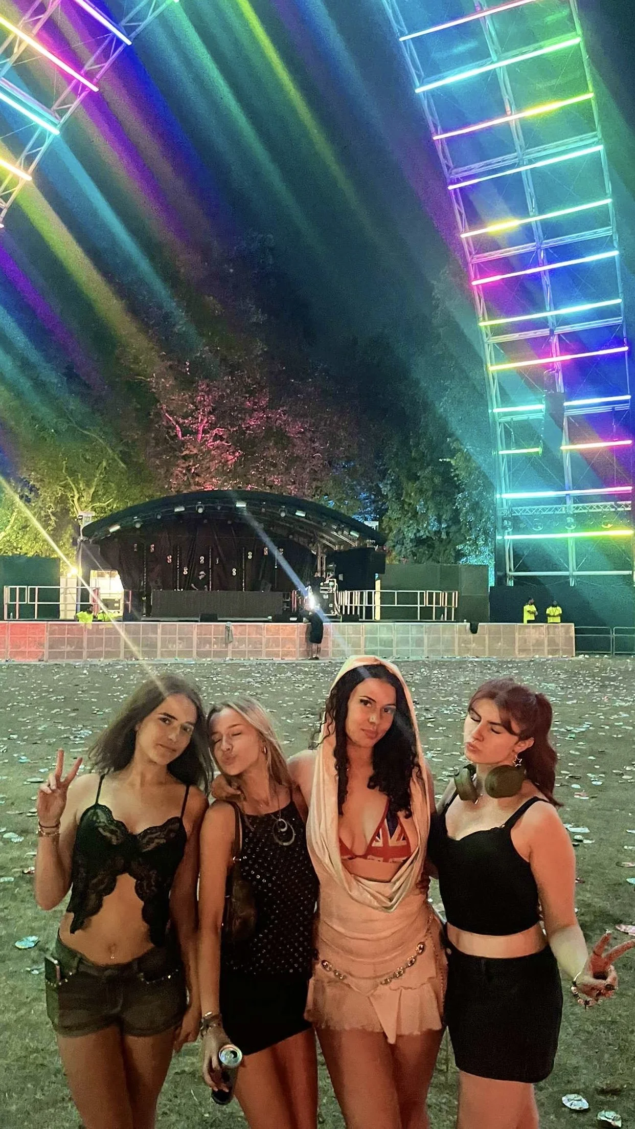 Four young women dressed in festival attire posing together under colorful neon lights at an outdoor music festival, with a stage in the background and trees illuminated by the lights.