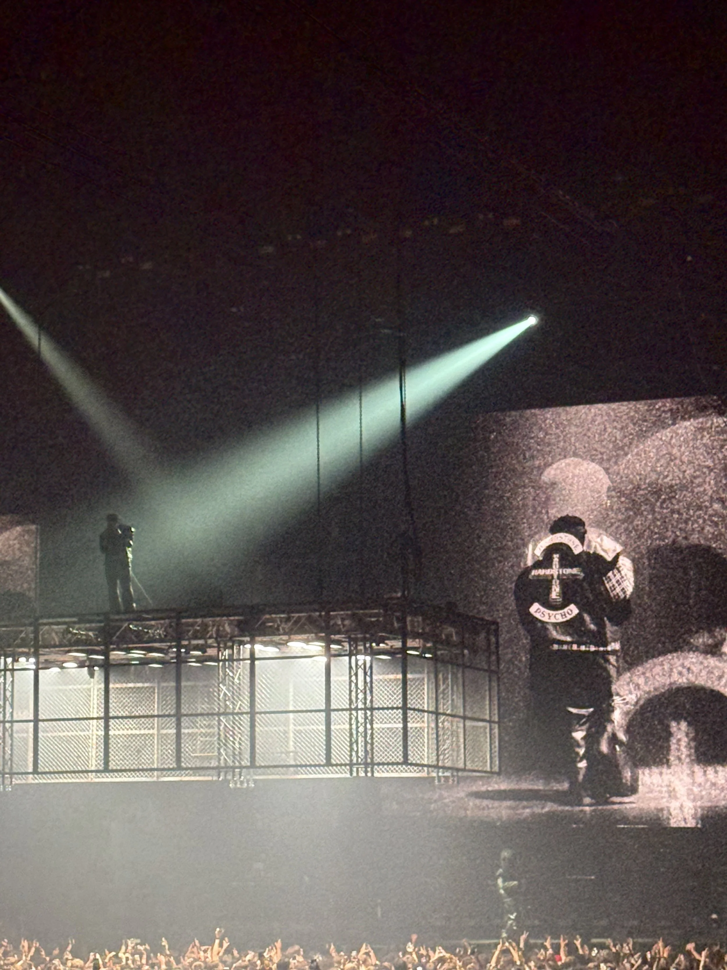 Stage with Don Toliver: standing on an elevated platform holding a microphone whilst projected on a big screen, wearing a jacket with an emblem reading "PSYCHO", crowd cheering at the bottom, dark background with a large screen displaying an image.