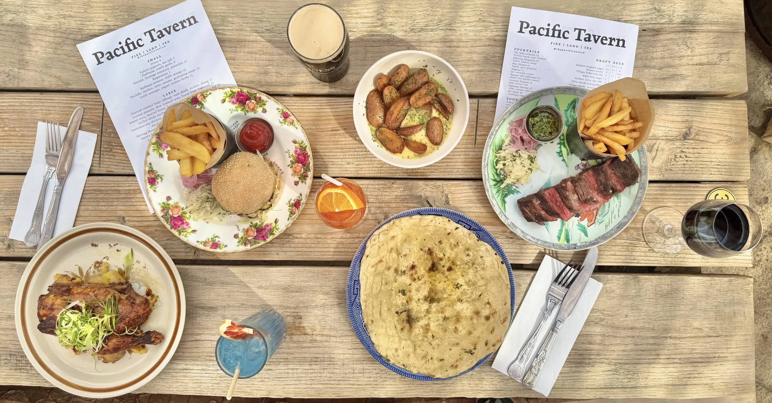 A wooden table with various dishes including a burger with fries, a steak with fries, grilled potatoes, a large piece of flatbread, a plate of ribs with fries, and glasses of beer, red and blue-colored drinks, and a glass of red wine, with menus from Pacific Tavern.