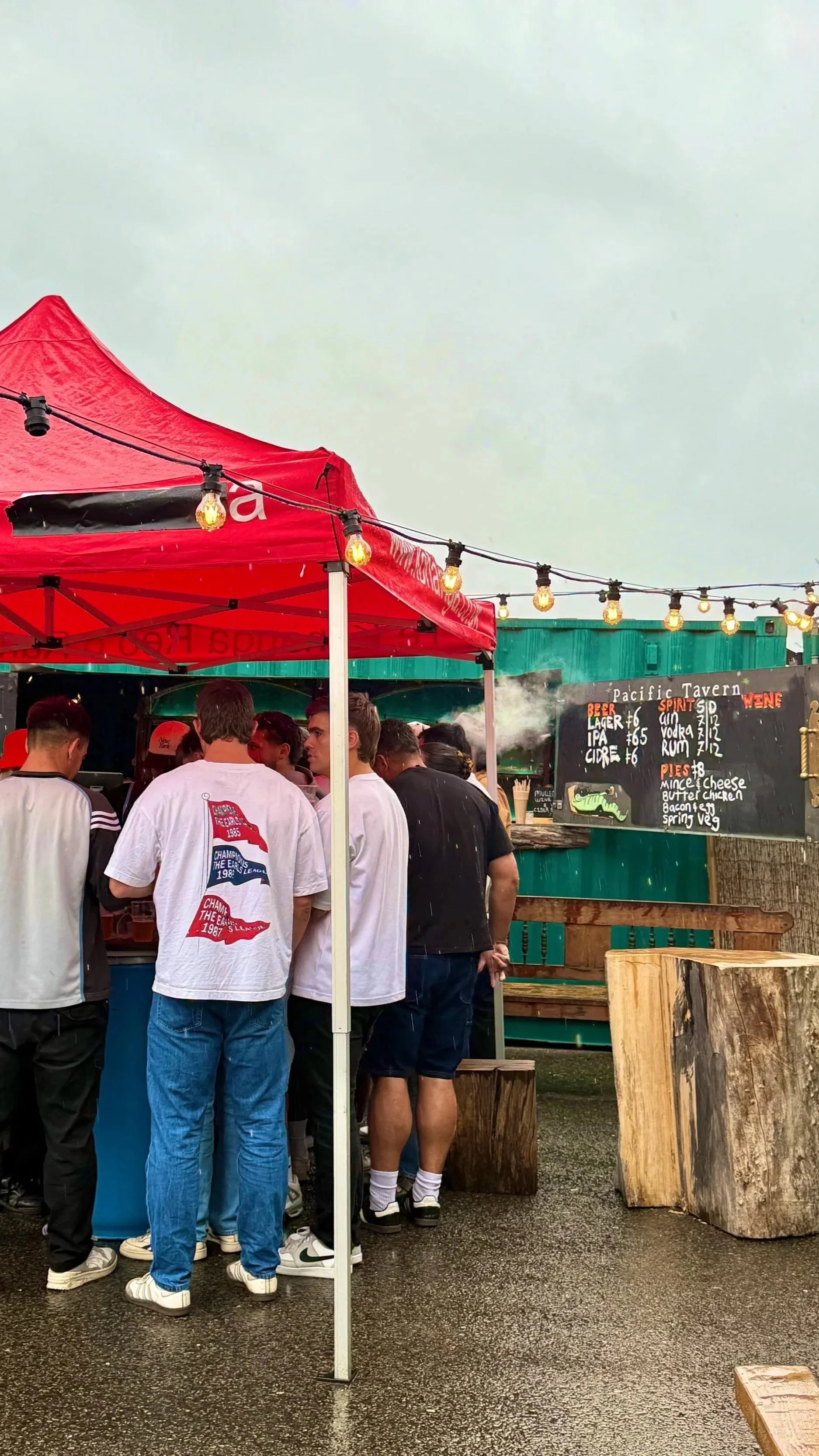 People waiting in line at an outdoor food stand with a red canopy, string lights overhead, and a menu board displaying drinks and pies, on a rainy day.