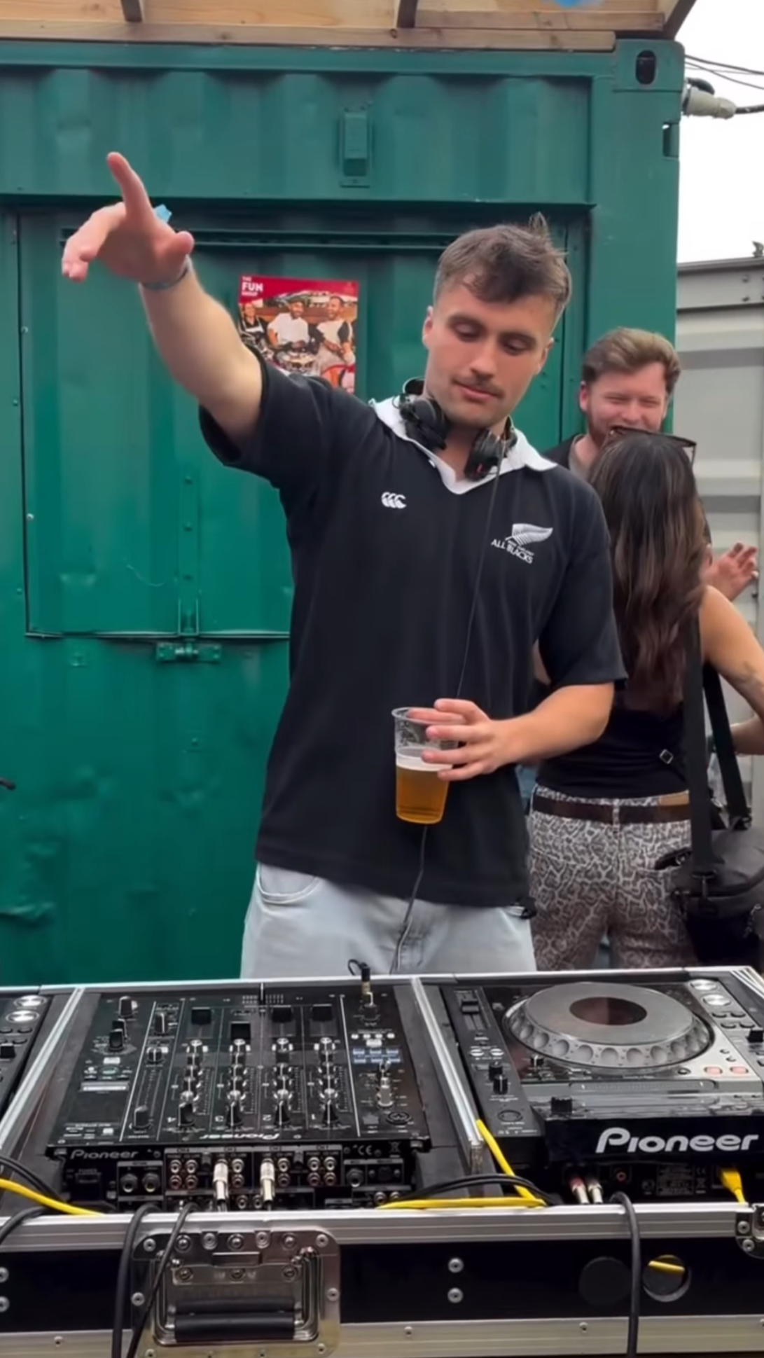A man with headphones around his neck is DJing at an outdoor event, holding a drink in his left hand while raising his right hand in the air. There are two other people smiling in the background, and a DJ setup with Pioneer equipment is in front of him.