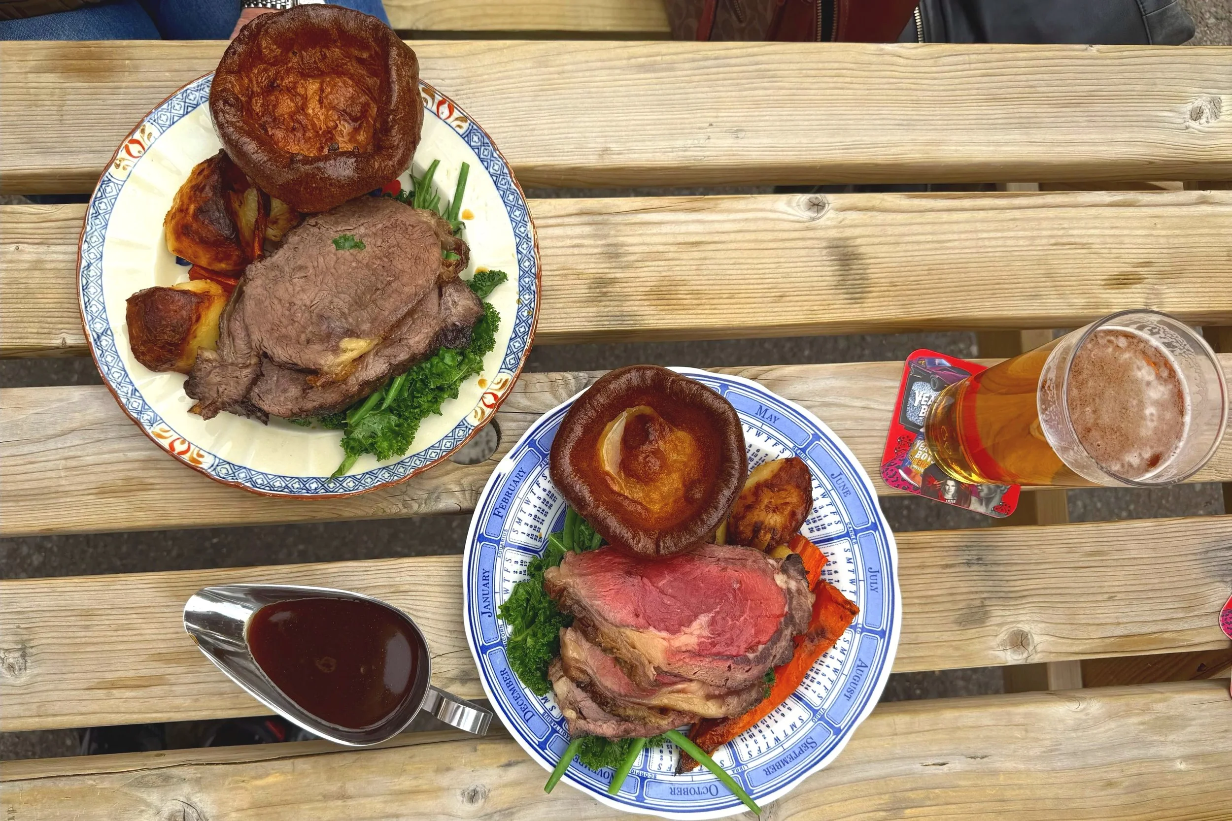 Two plates of beef dinner with Yorkshire pudding, roasted vegetables, and gravy, along with a pint of beer and a gravy boat, on a wooden picnic table.