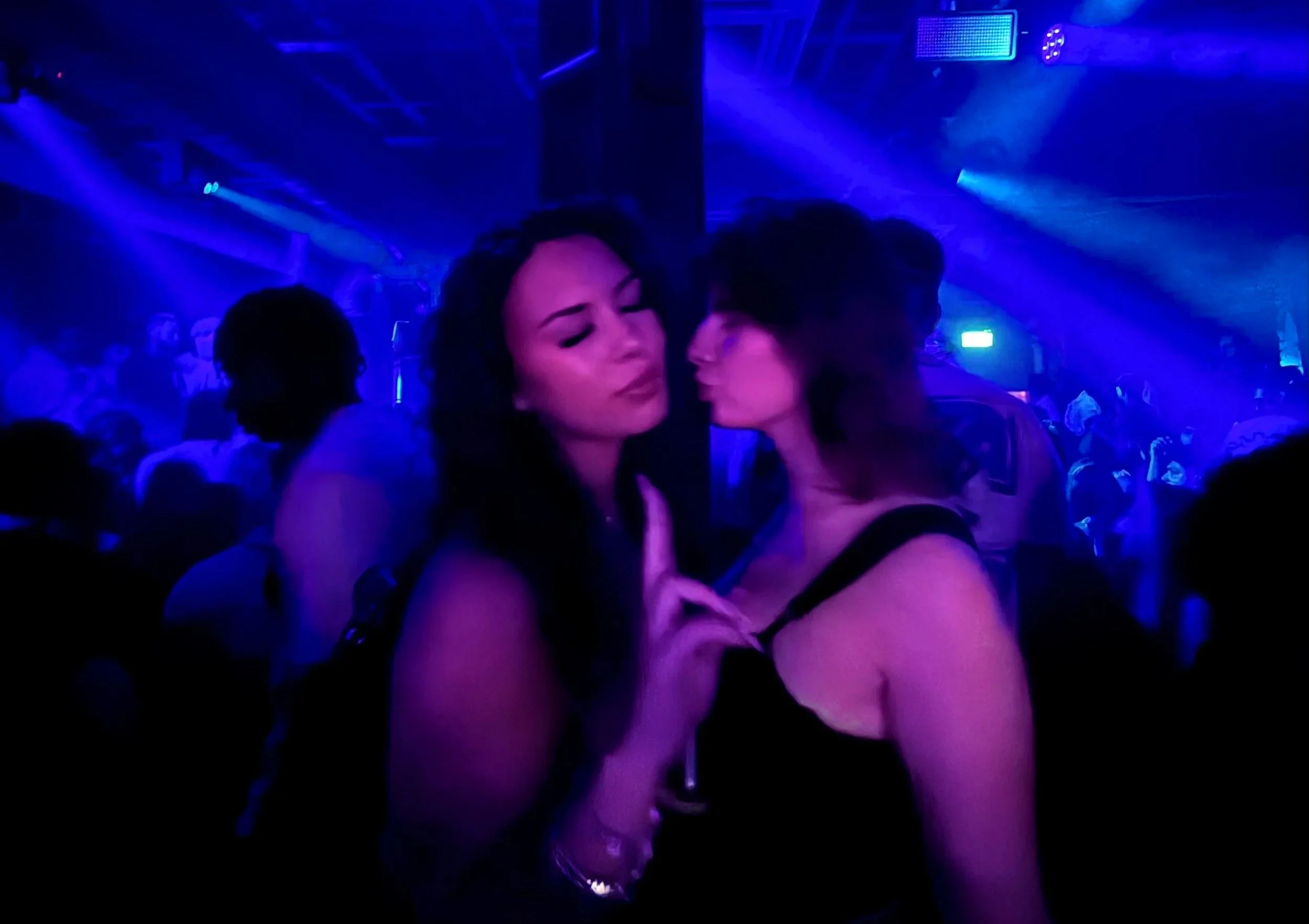 Two women in a nightclub with blue and purple lighting, one is making a peace sign, and the other is leaning in for a kiss. XOYO London Club