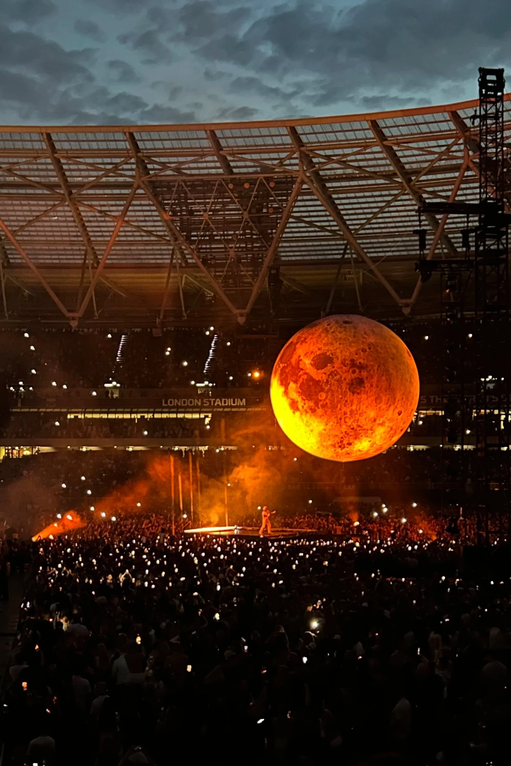 A large audience in a stadium watching a performance on stage with a giant, glowing moon prop hanging above. The Weeknd London Stadium