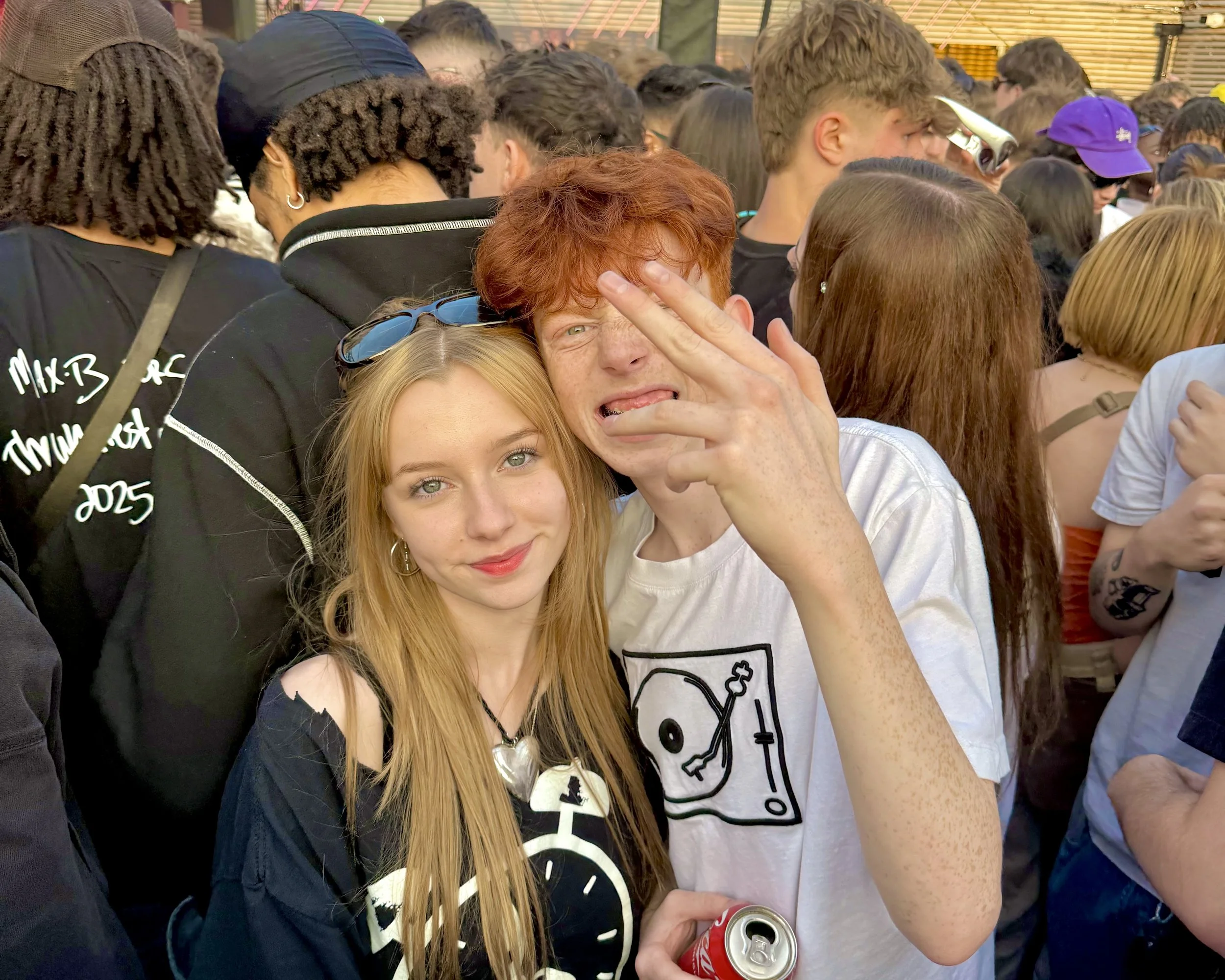 A large crowd of people standing closely together, with some people holding cans of soda. Two women are prominently in the foreground, one with red hair and freckles making a peace sign over her face and the other with blonde hair looking directly at the camera.