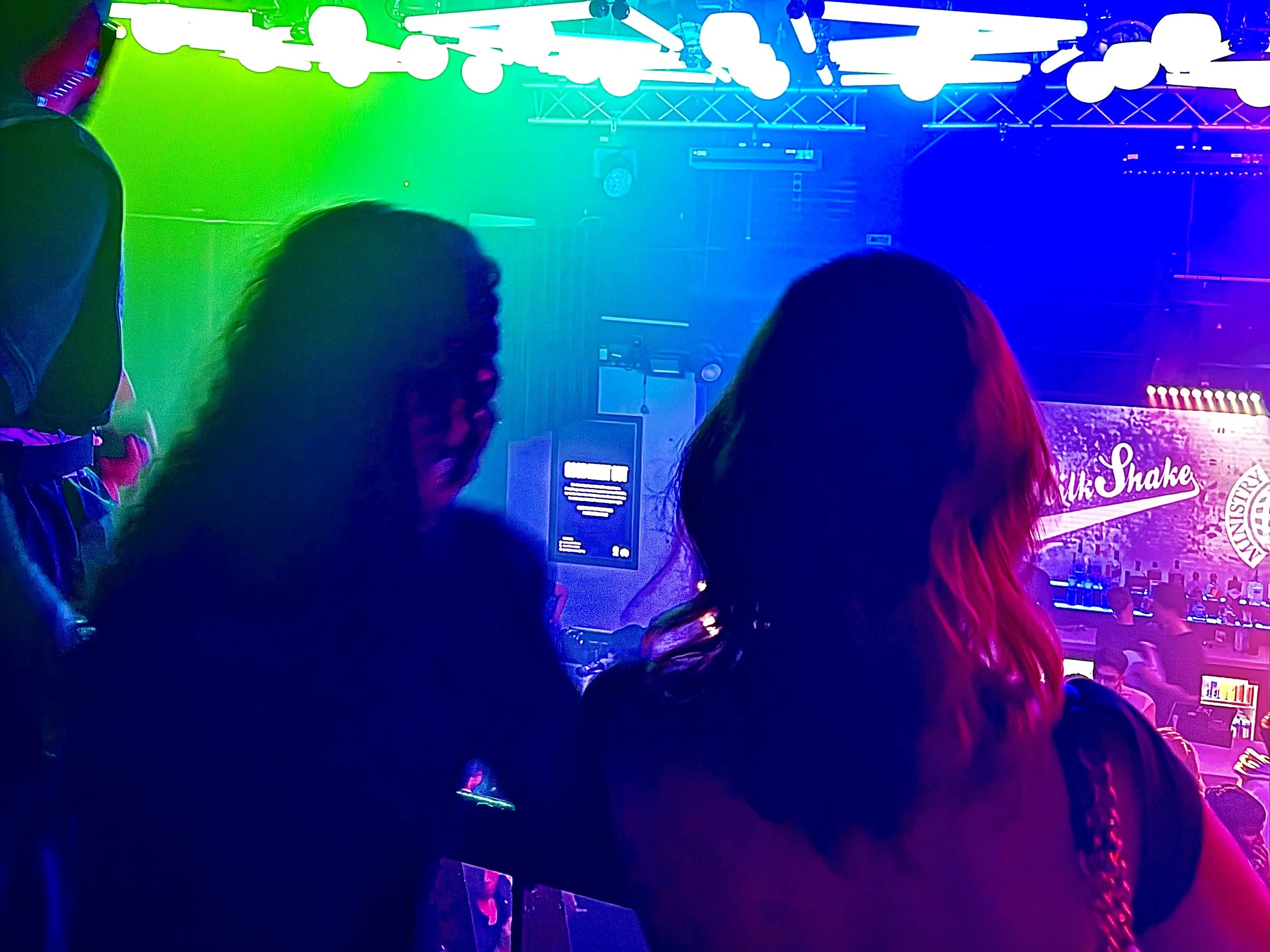 Two women with long hair in a nightclub or concert venue, illuminated by green, purple, and blue stage lights, with a stage in the background displaying a logo and people sitting at tables. Ministry Of Sound
