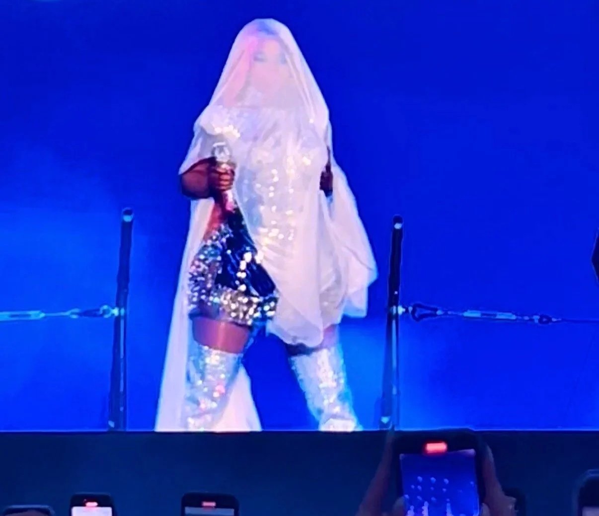Person dressed in a shiny, sparkly outfit with thigh-high boots, partially covered in a sheer cape, standing on stage with a dark background.