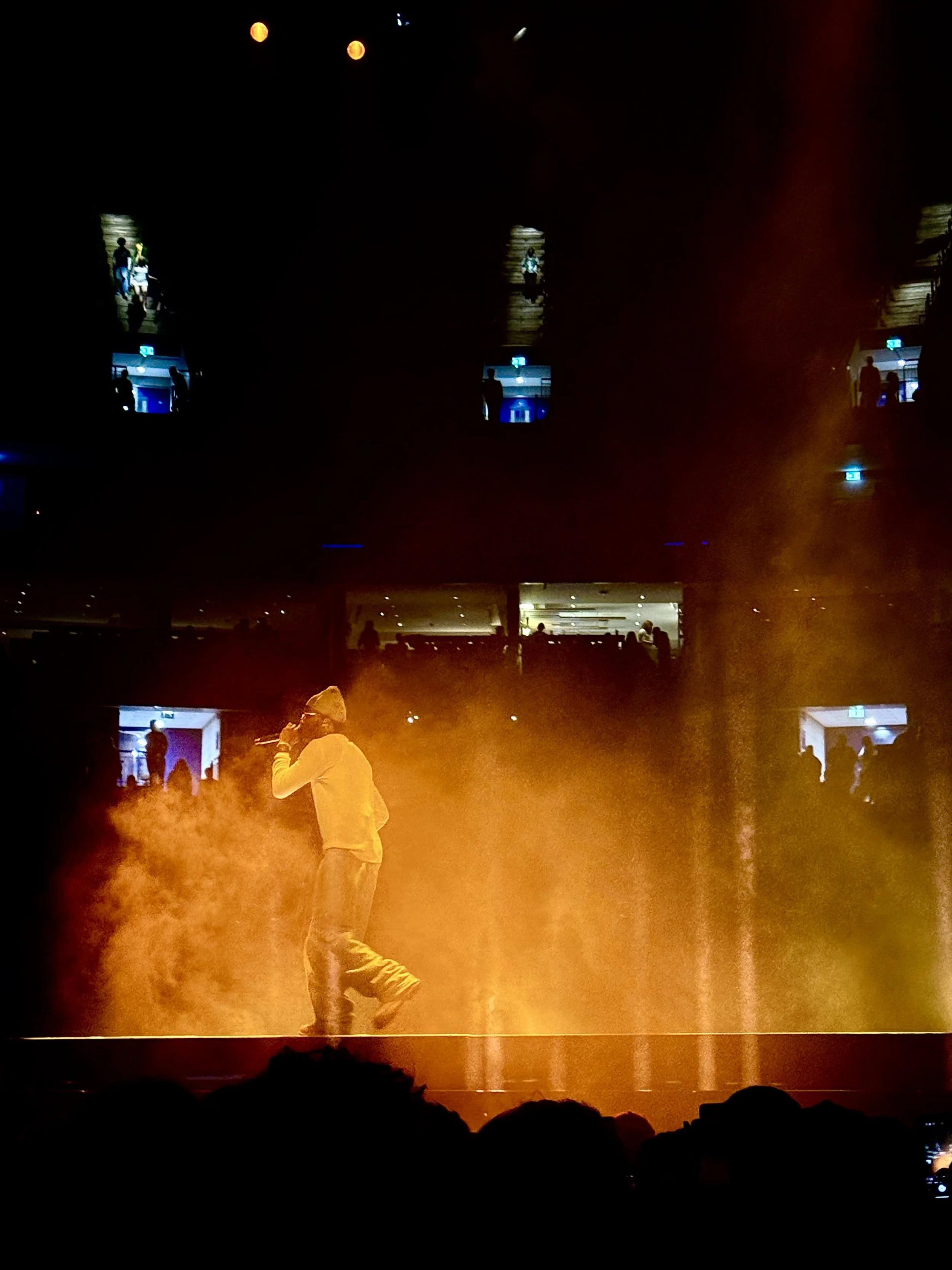 A performer onstage singing into a microphone with orange stage lighting and smoke effects, and audience members visible in the foreground. Lil Yachty O2 Arena London