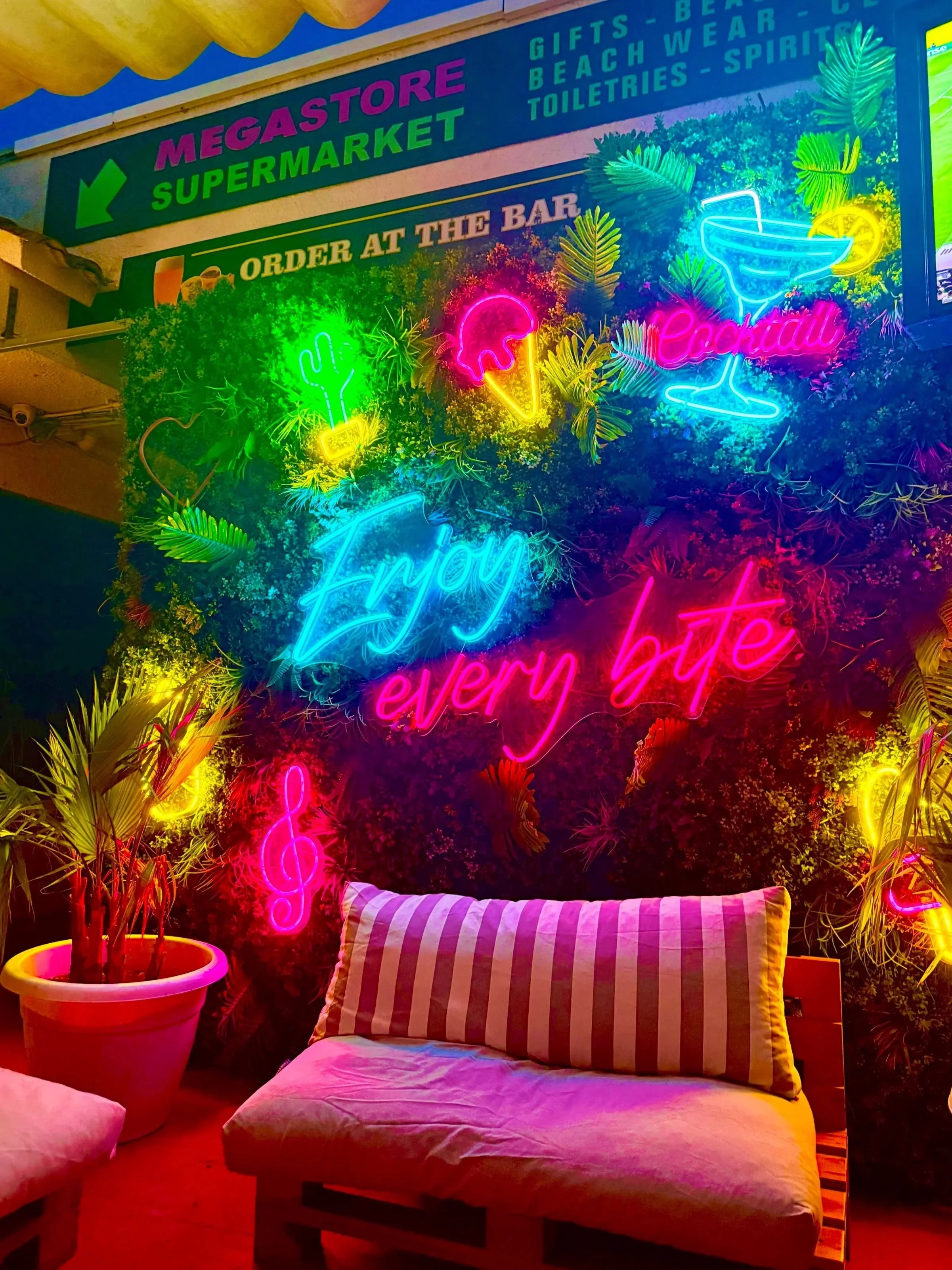 Neon signs with tropical leaves, ice cream, cocktail, cactus, and music notes on a green leafy wall, with a striped cushion on a bench and a potted plant in the foreground, at a colorful bar or lounge area.