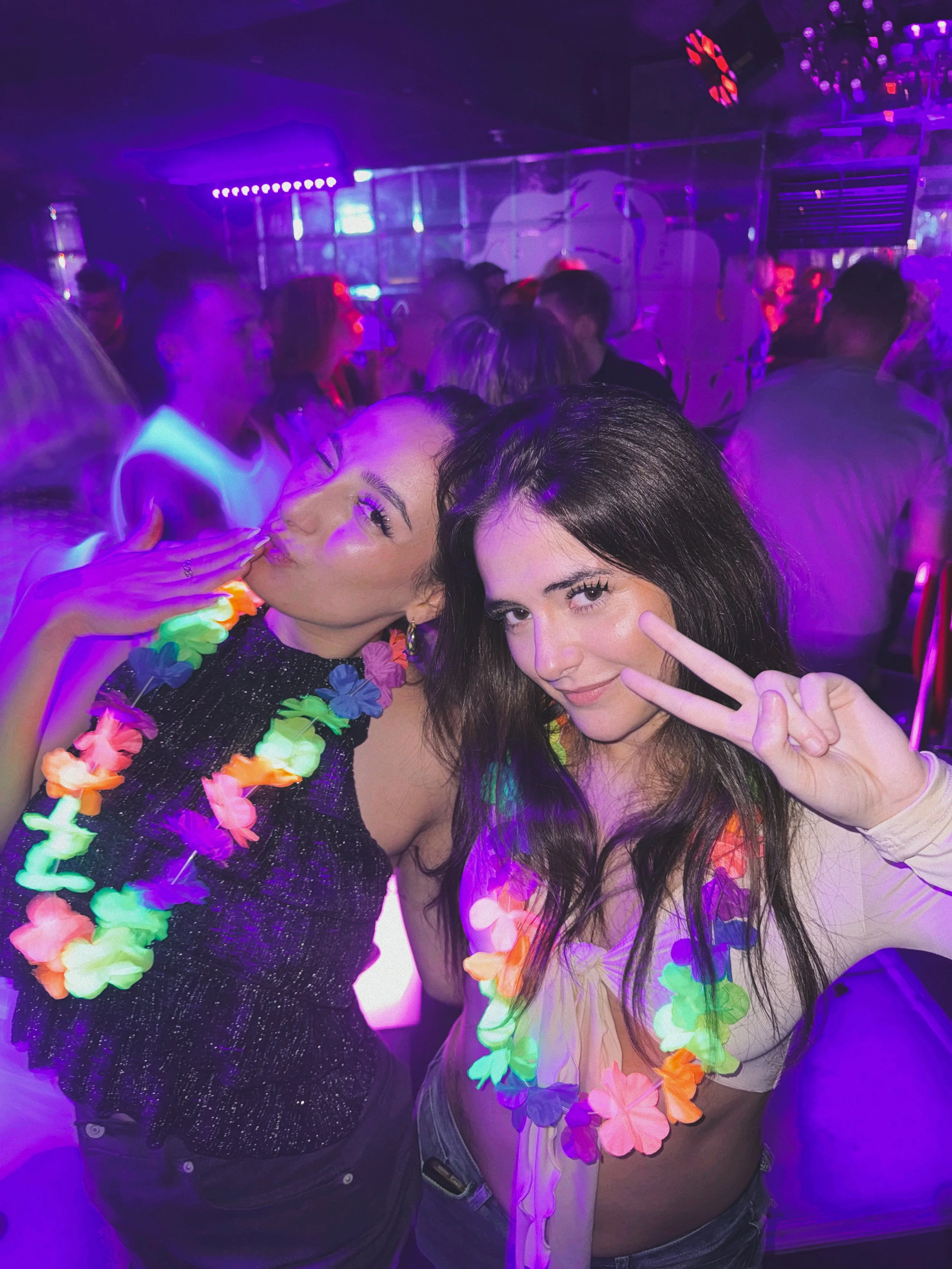 Two women are at a party with purple and neon lighting. They are making peace signs and smiling. One woman has dark brown hair and is wearing a white shirt, the other has dark hair and is wearing a black sparkly top with a colorful flower lei.