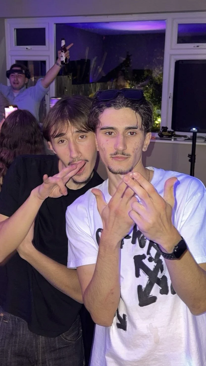 Two young men taking a selfie at a party, with a man in the background holding a beer and raising his arm in celebration. The photo is taken indoors during the evening.