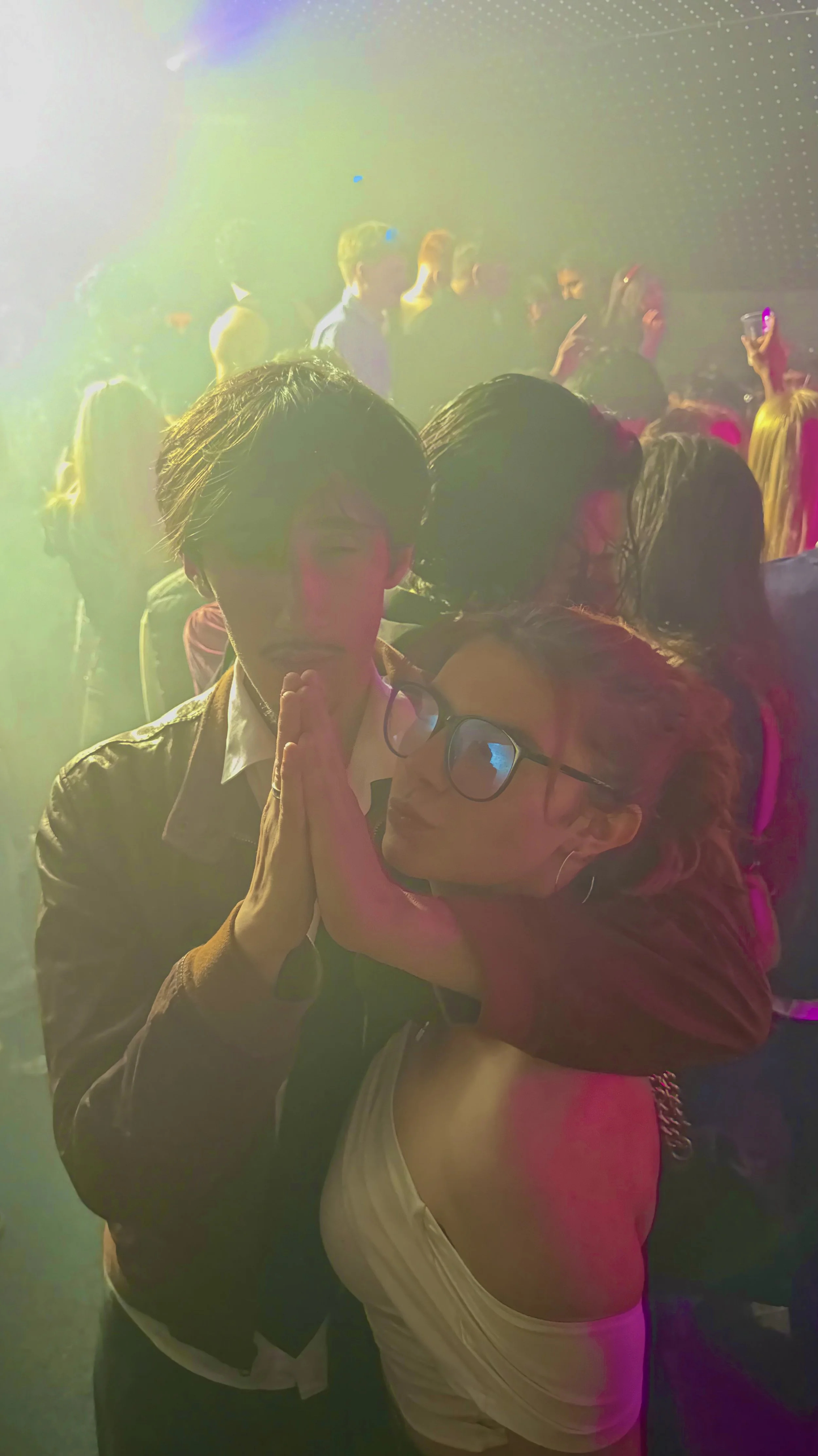 A man and a woman with glasses, standing close together with their hands pressed together in a praying pose at a crowded club or club with colorful lighting. Lightbox London