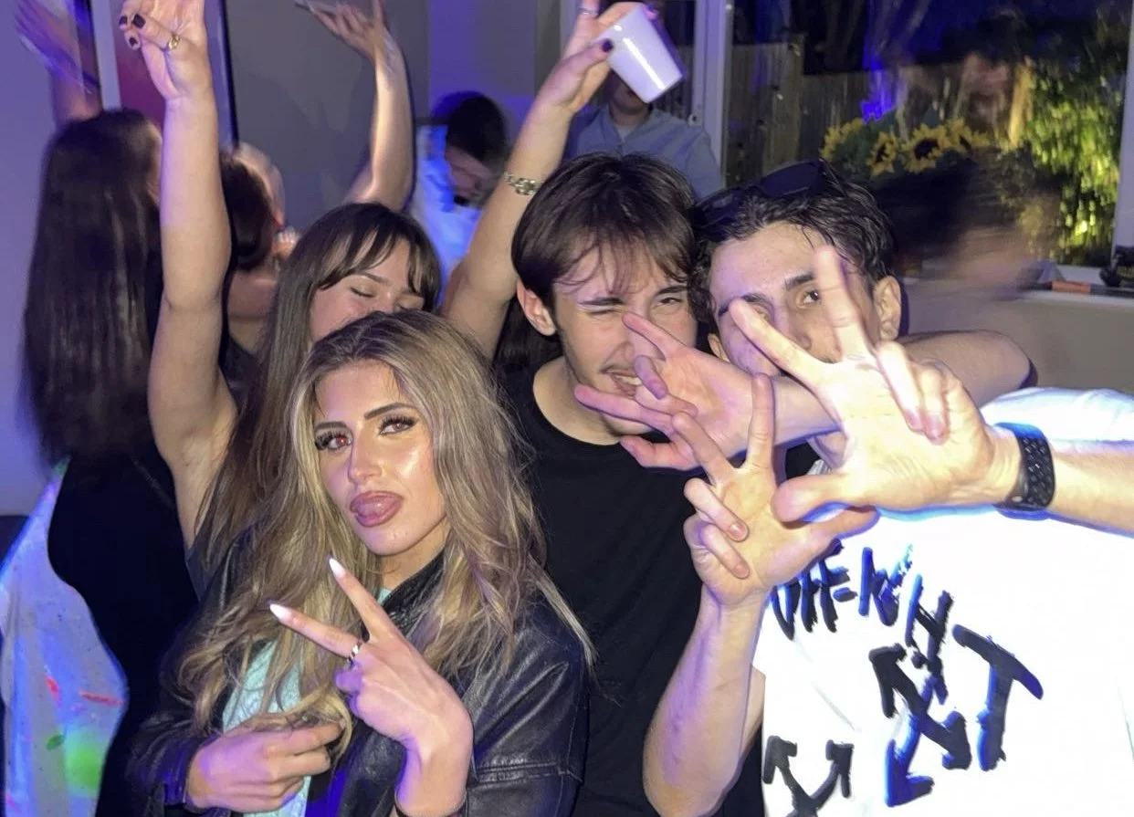 Group of young adults enjoying a party, with some making peace signs and playful gestures, in an indoor setting with colorful lighting.