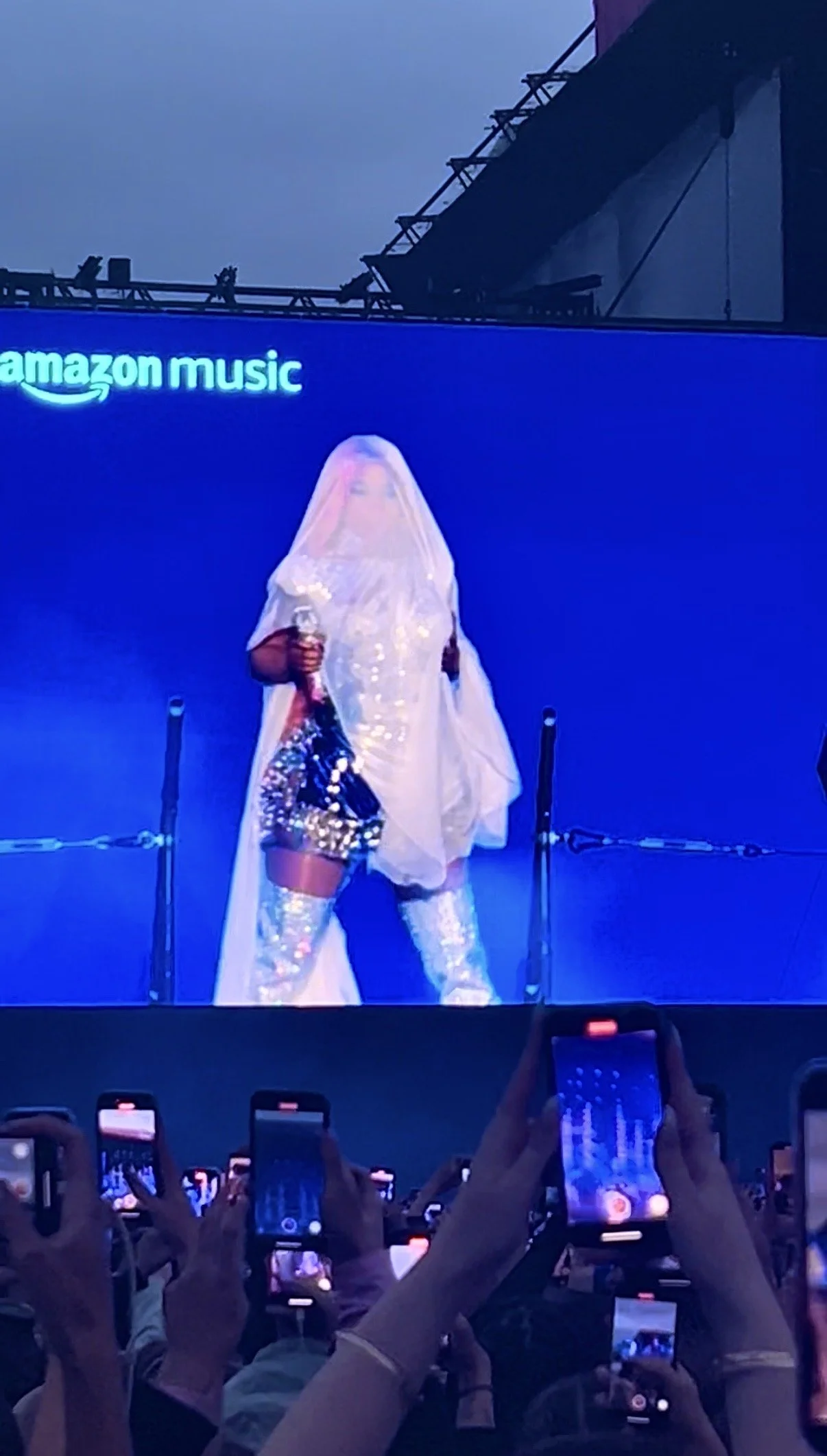 A performer on stage during a concert, dressed in sequined shorts, thigh-high boots, and a flowing veil, holding a microphone, with many audience members taking photos or videos. Nicki Minaj Wireless Festival 2024