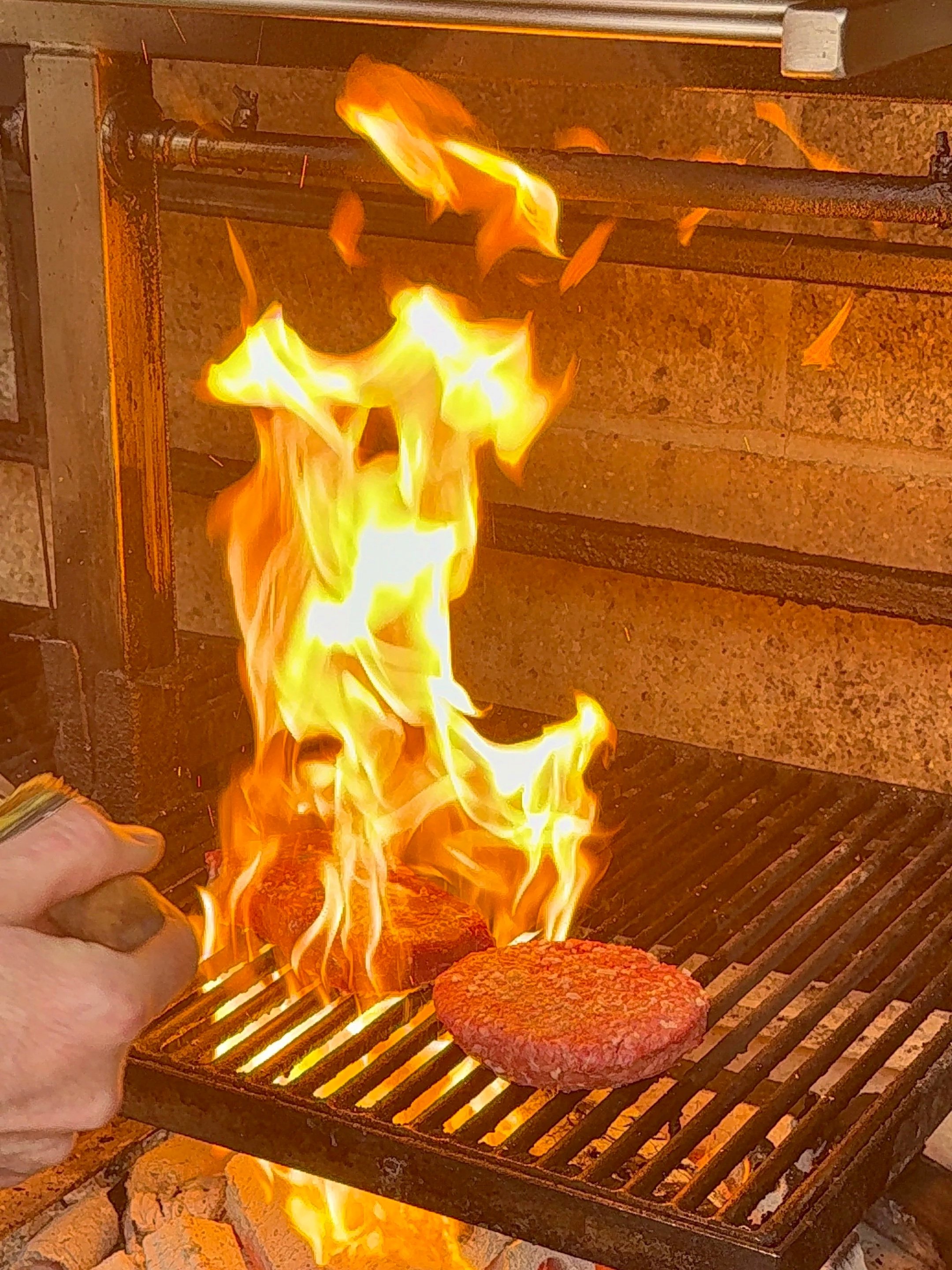 A person is grilling hamburger patties on a barbecue grill with flames rising from the cooking surface.