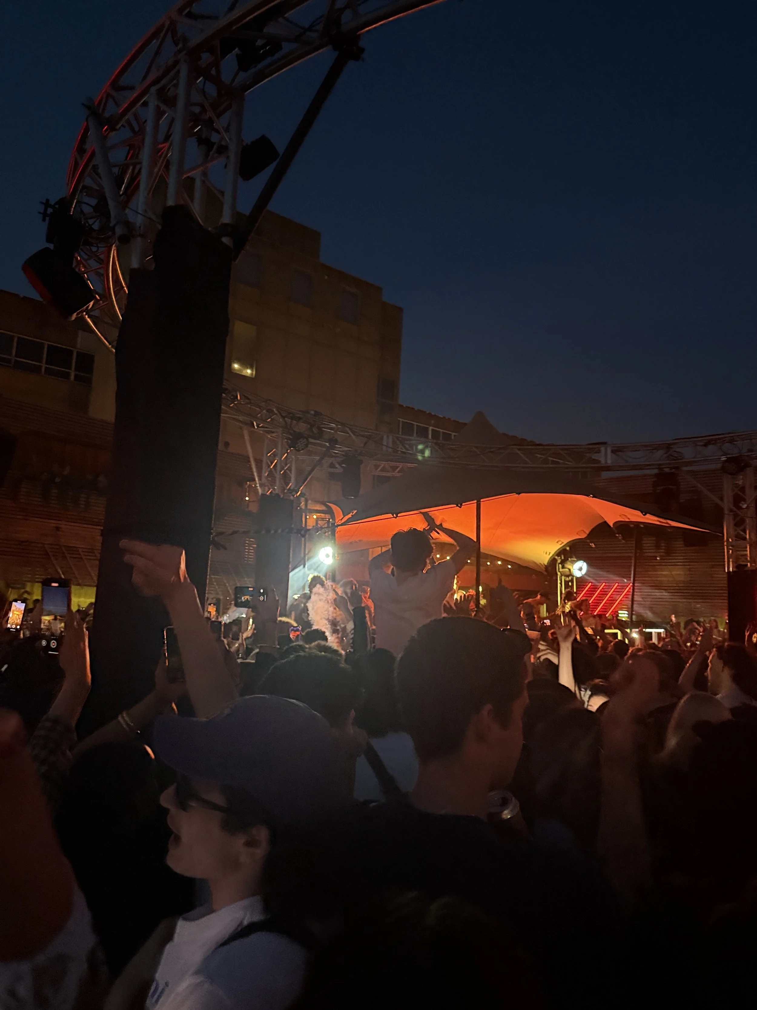 A crowd of people at an outdoor concert during the night, with some raising their hands and taking photos. There is a stage with orange lighting and a band performing, and the sky is dark.