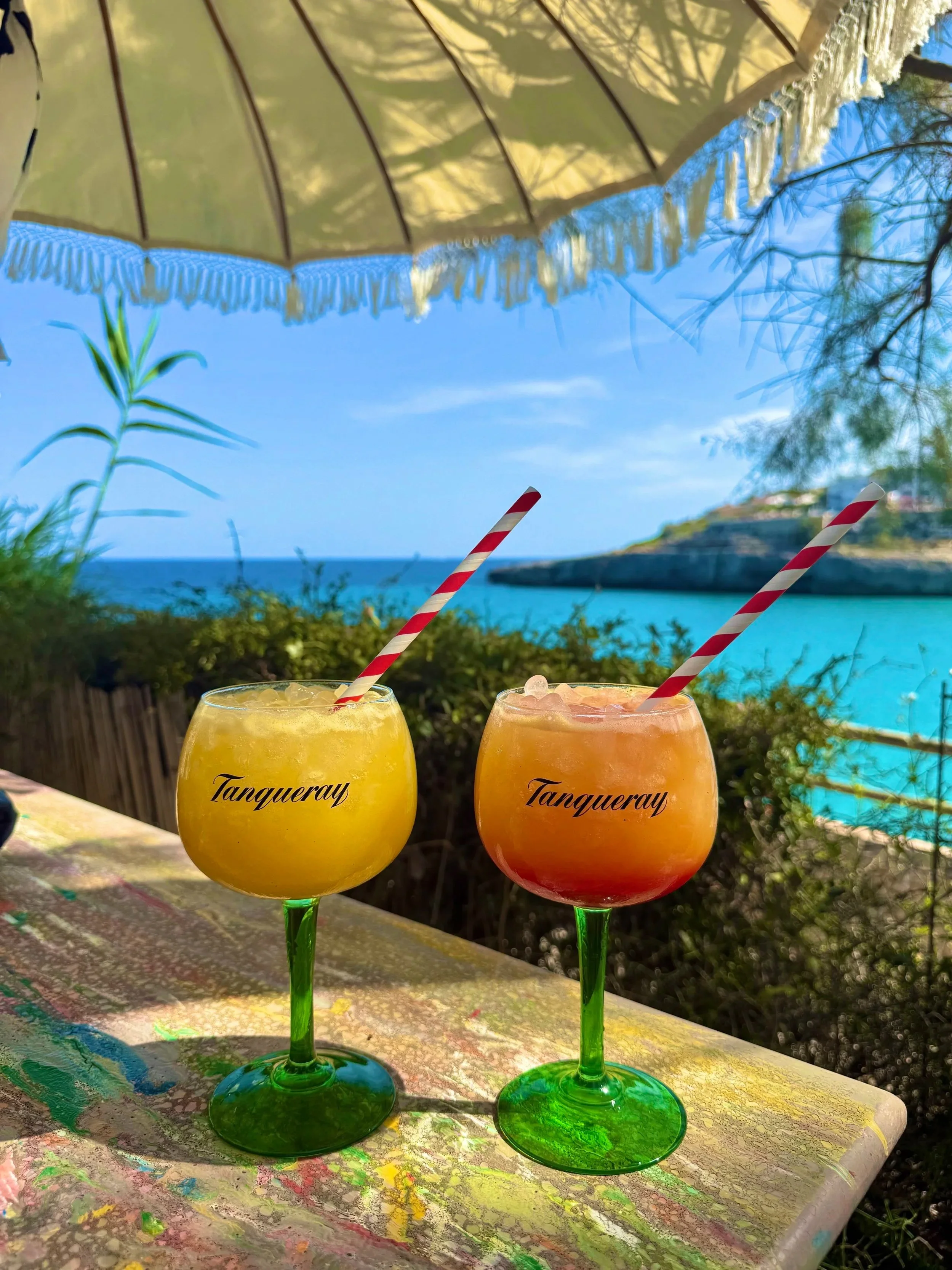 Two colorful tropical drinks in cocktail glasses with red and white striped straws, set on a paint-splattered table under a large umbrella, overlooking a beach with turquoise water and a rocky shoreline, with greenery in the foreground. Majorca