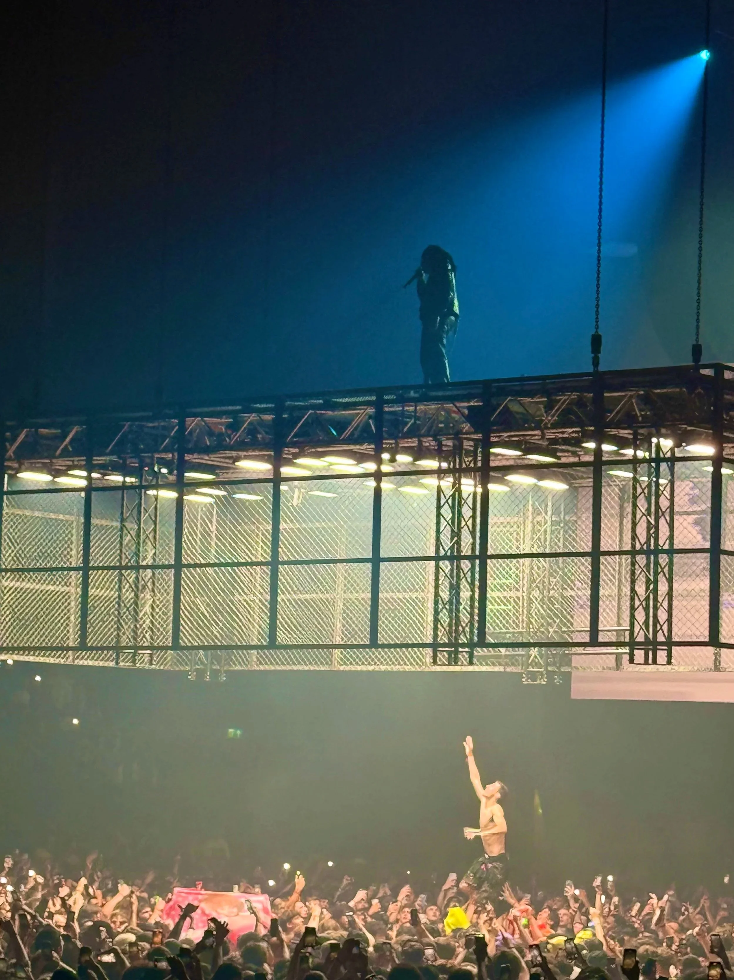 A concert crowd with a performer on the ground reaching upward and a person on a raised platform above, singing into a microphone surrounded by stage lighting and structures. Don Toliver