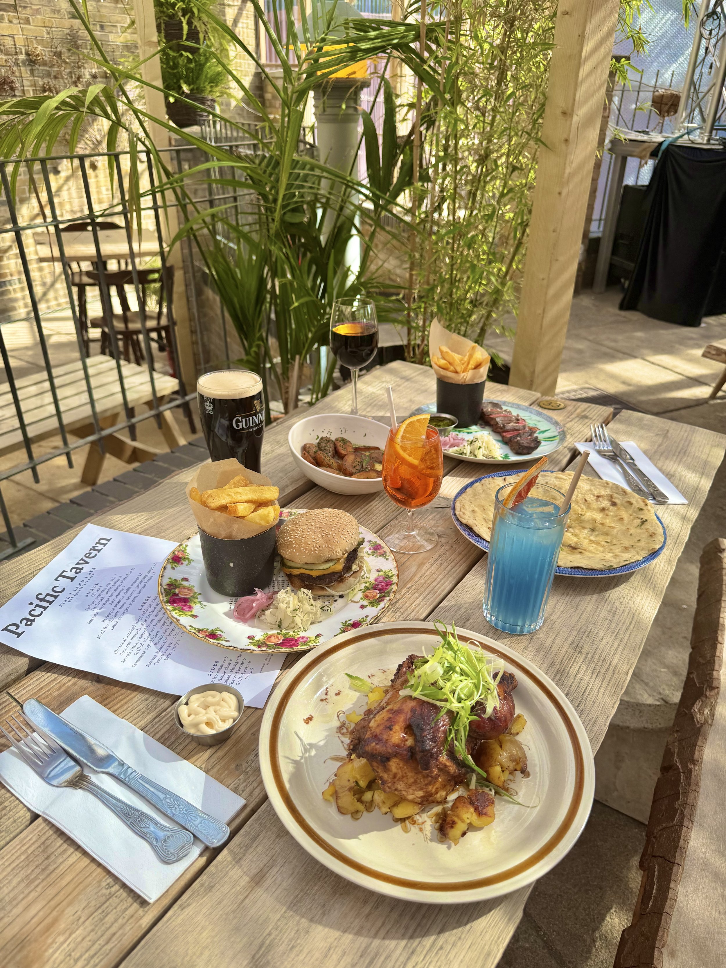 A table set for a meal at a restaurant with various dishes including a burger, ribs, fries, chips, bread, sausages, and drinks such as beer, wine, and colorful cocktails, in an outdoor setting with plants and natural lighting.