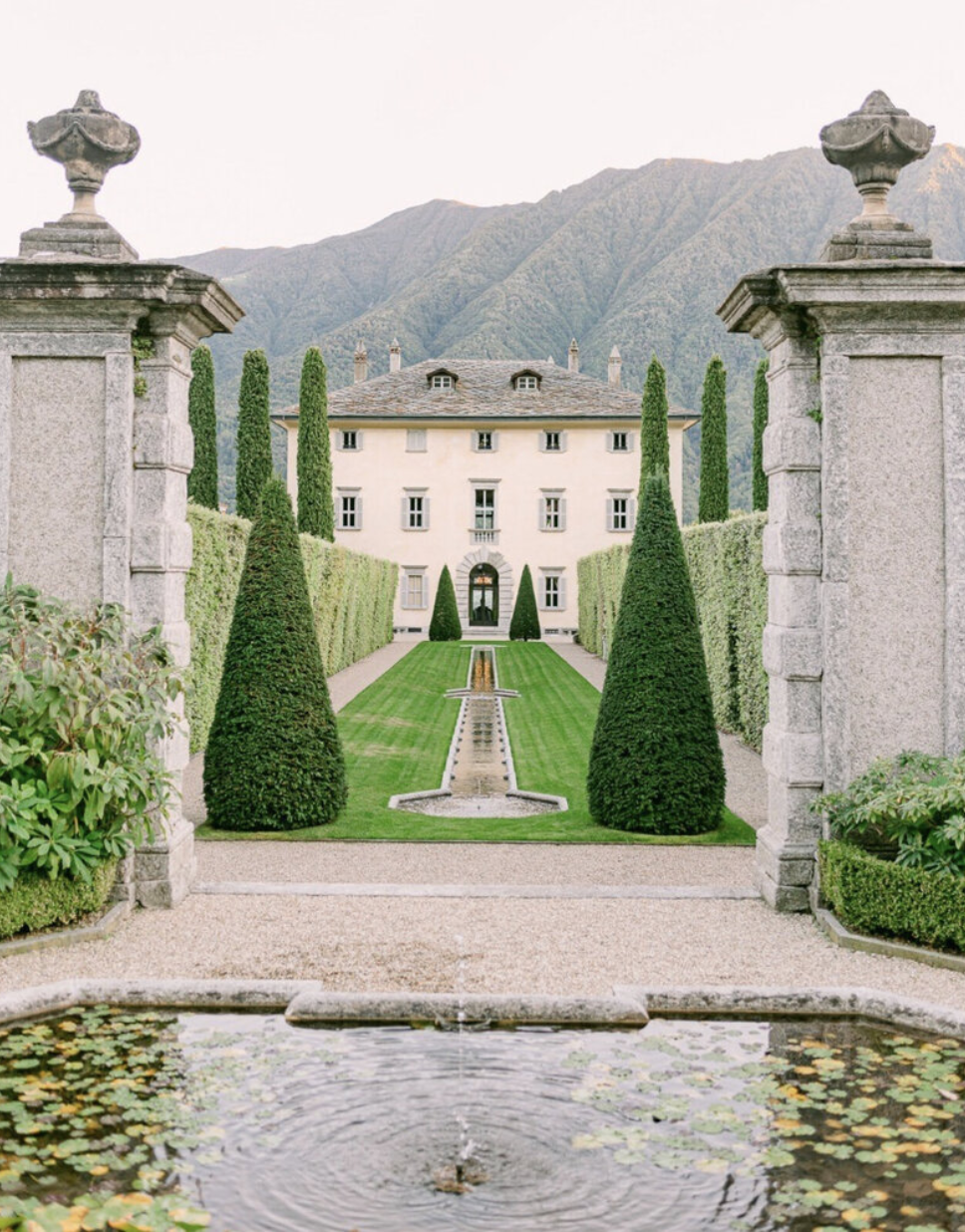 Villa balbiano Lake Como A view of a grand estate with a manicured garden featuring a fountain, symmetrical topiary, and tall cypress trees, with a large house and mountains in the background.