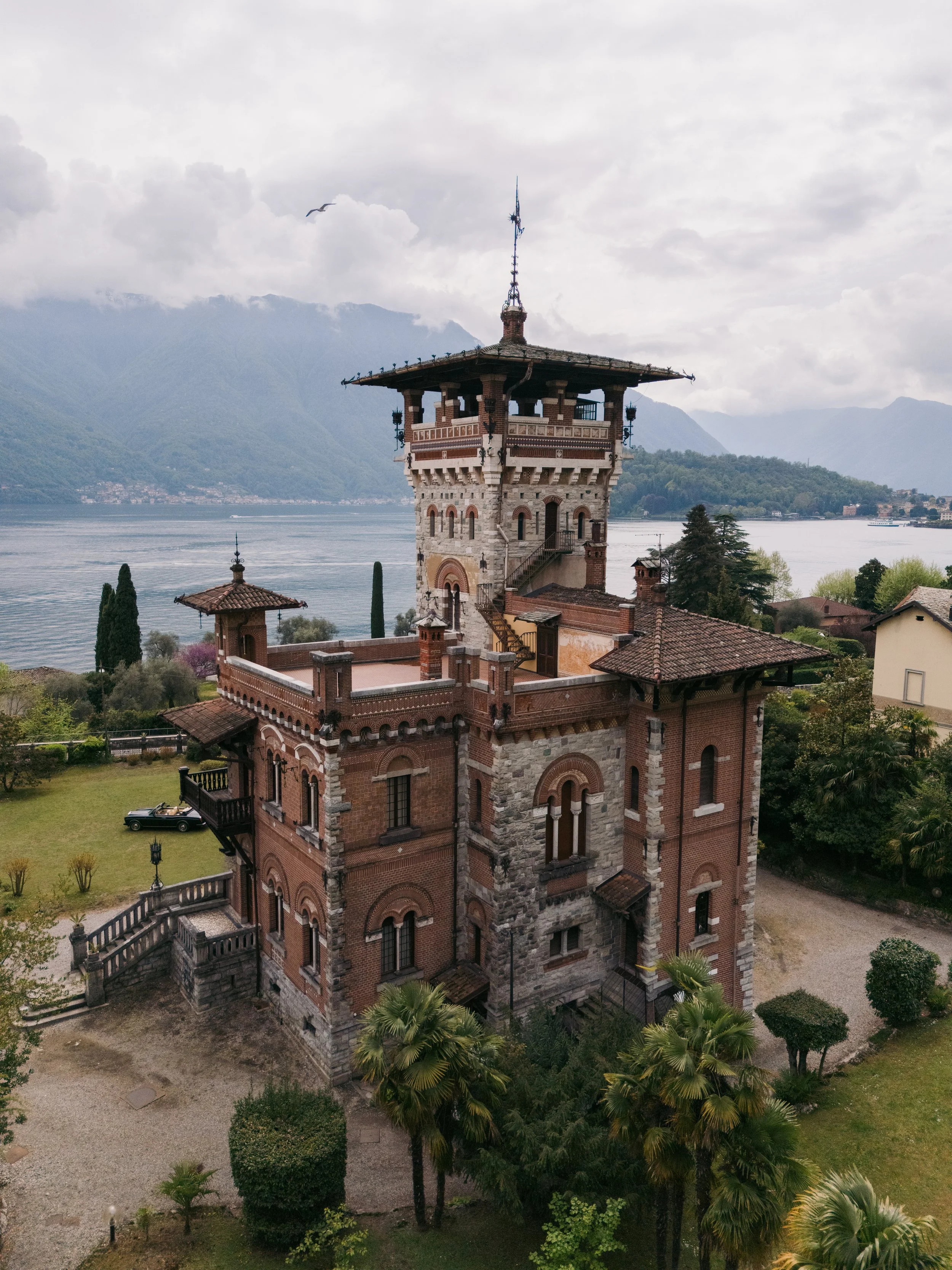 Villa Pessina A tall, historic brick and stone castle with a distinctive tower, situated near a lake with mountains in the background, under a cloudy sky.