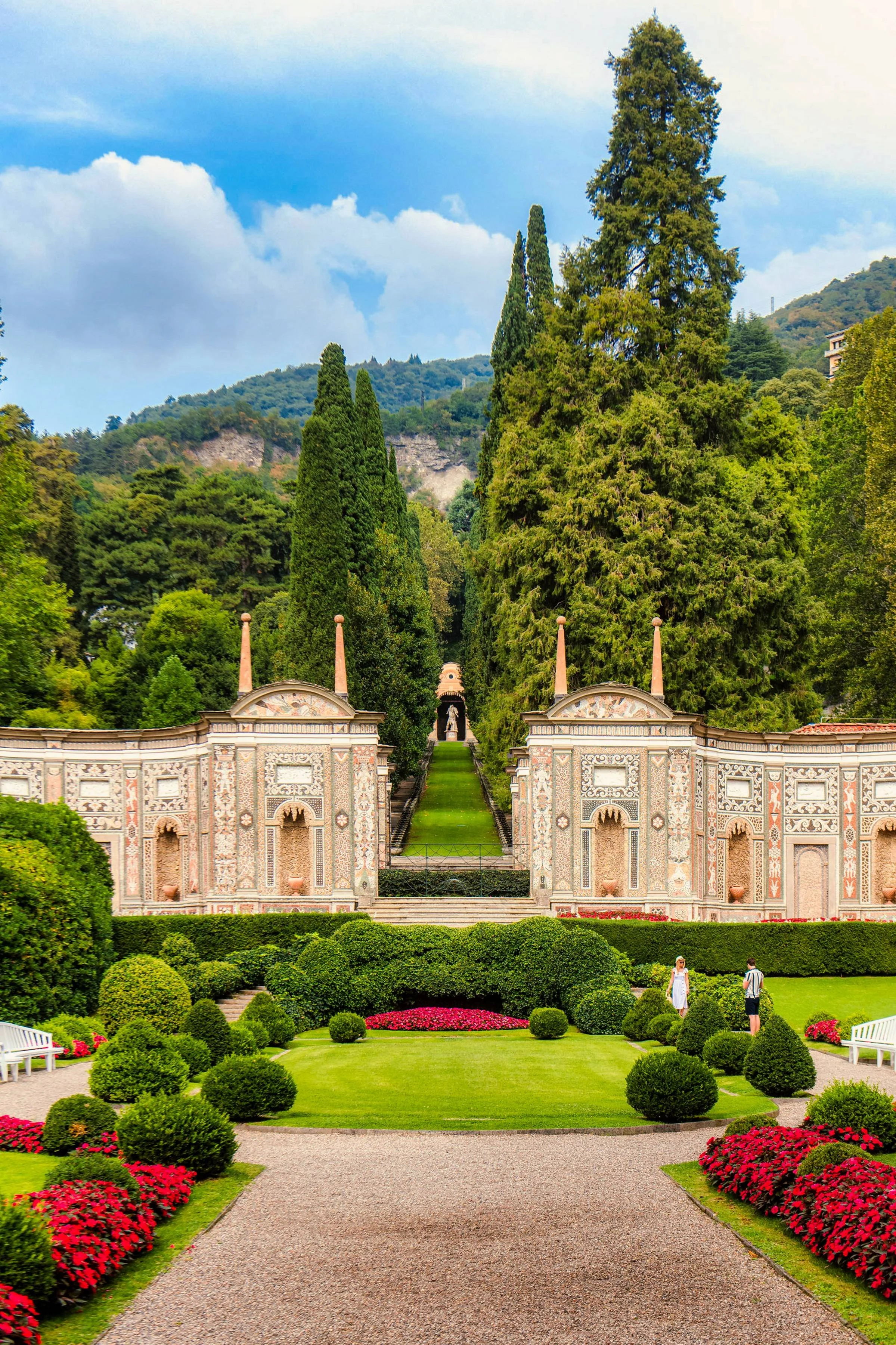 Villa d'Este garden with lush green grass, trimmed bushes, and vibrant red flowers, leading to a historical building with ornate architecture and a statuesque figure on a pathway, surrounded by tall trees and scenic hills in the background.